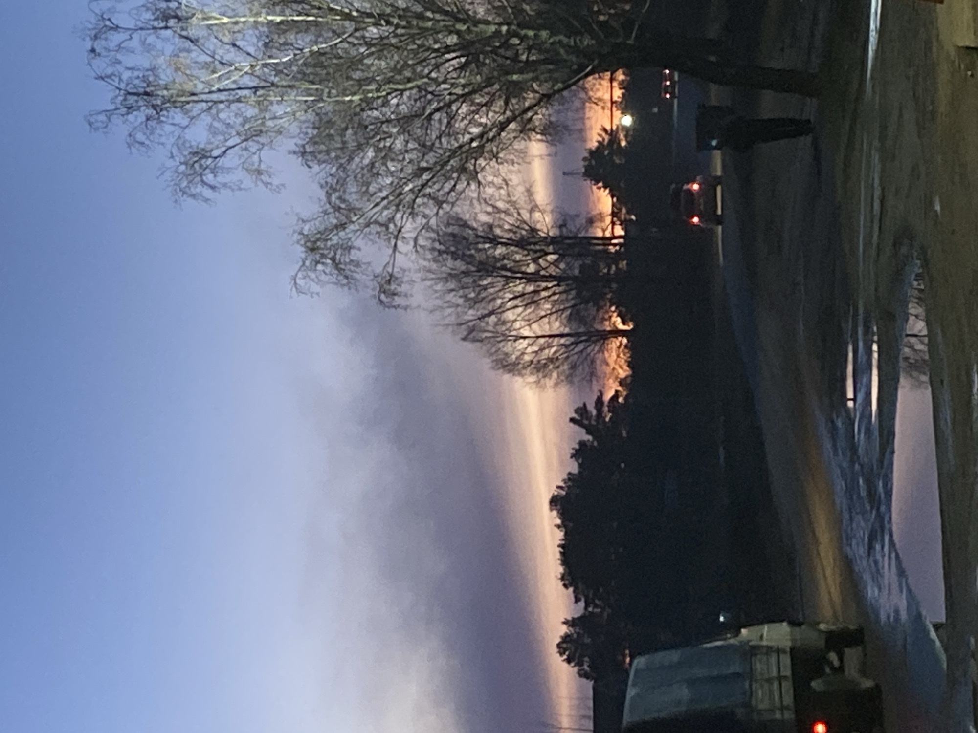 The image depicts a twilight scene in Noviy, Russia. The sky exhibits a gradient of colors, transitioning from blue to orange. Silhouetted trees line the horizon. A vehicle with illuminated tail lights is visible on the road. Water is standing on the roadside and there are street lights.
