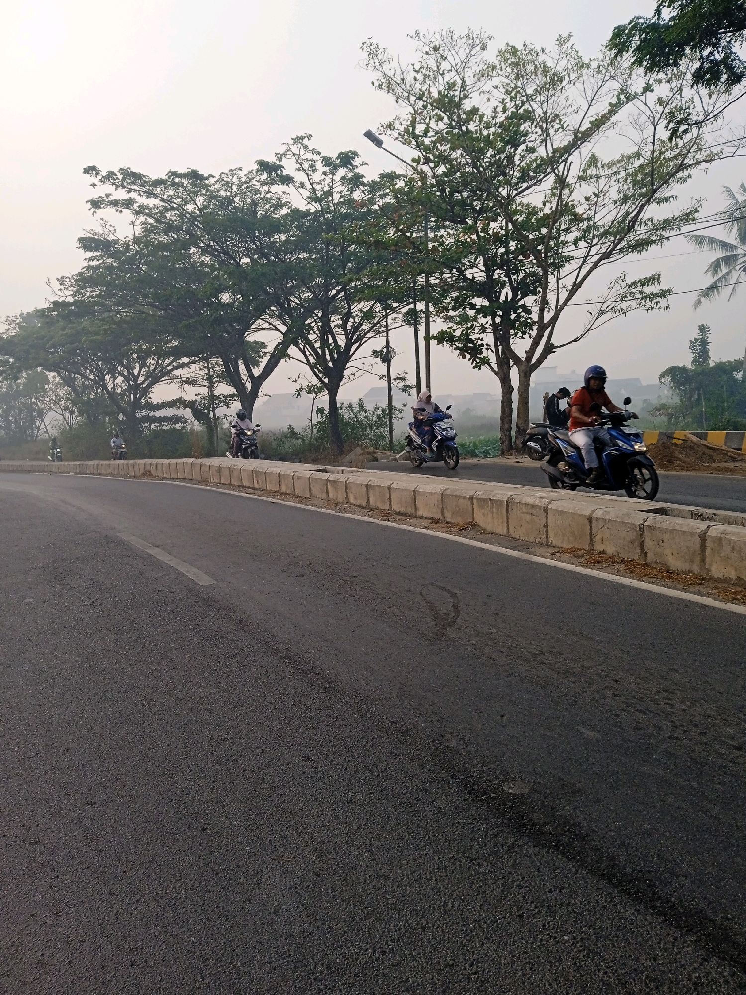 The image depicts a paved road in the city of Banjarmasin, Indonesia, lined with concrete barriers and trees. The sky is overcast, suggesting a hazy or humid atmosphere. Several individuals are riding motorcycles on the road. On the right, a person wearing a red shirt and a blue helmet is riding a blue motorcycle. To the left, another person wearing a headscarf is riding a blue motorcycle. Further in the distance, more motorcycles are visible. The road appears to be a main thoroughfare with vegetation and some buildings in the background. The overall scene conveys a typical urban or suburban streetscape in Indonesia.