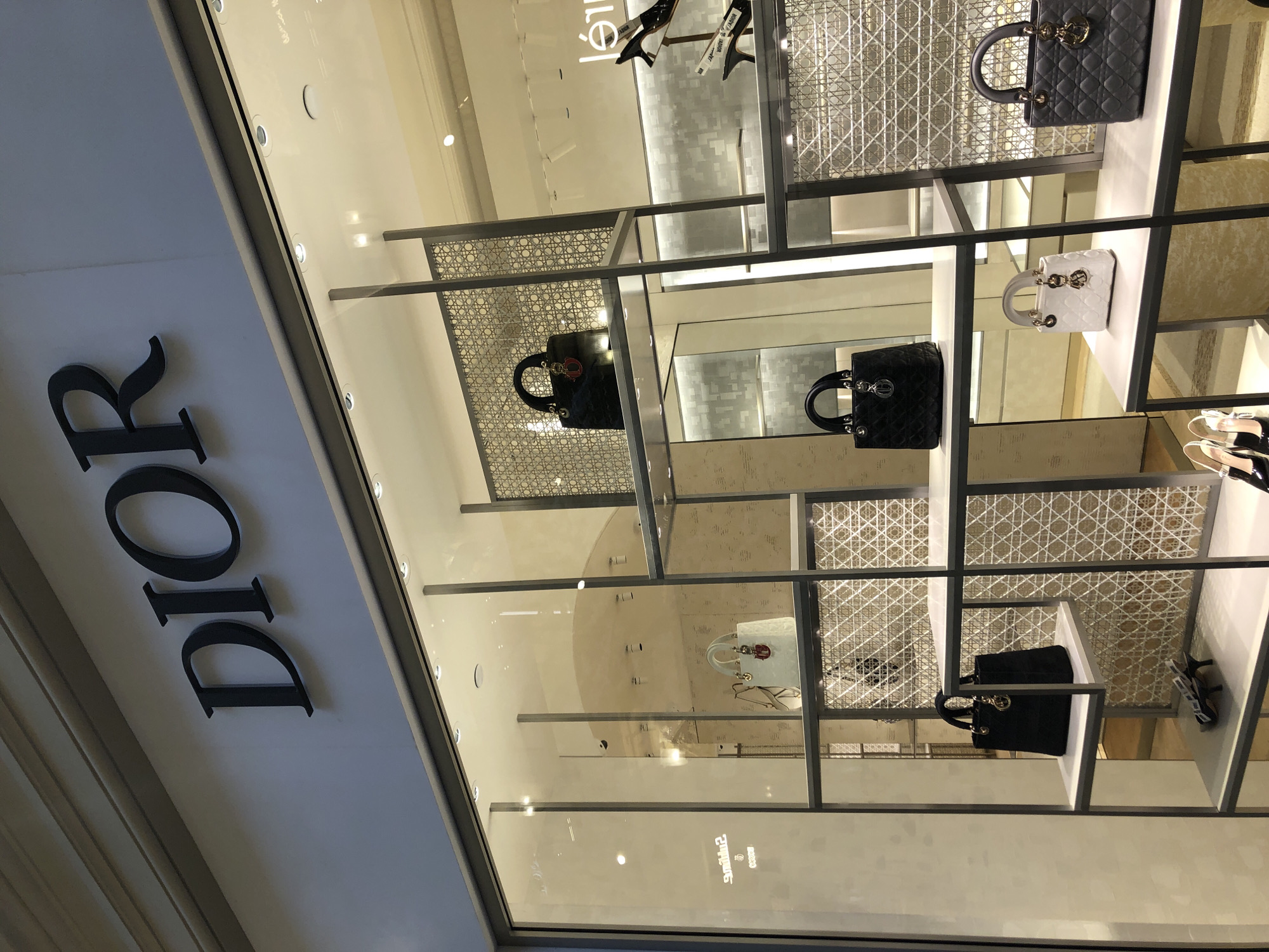 A storefront in Moscow, Russia, displays the "DIOR" brand name in bold black lettering on a white exterior wall. Behind a large glass window, a series of shelves showcase handbags of various styles and colors, including black and white quilted designs. Woven pattern backgrounds add visual texture. Shoes are also displayed on the shelves. The interior is brightly lit.