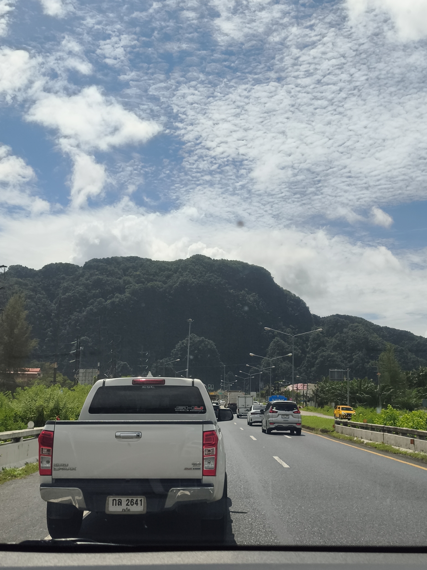 The image depicts a view from inside a vehicle traveling on a highway in Phang-nga, Thailand. The foreground is dominated by the rear of a white pickup truck, its license plate reading "กล 2641" and Thai characters below. The highway stretches ahead, with multiple lanes of traffic. Other vehicles are visible, including a silver SUV in the middle lane, a white truck, and a yellow vehicle further down the road. The roadside is lined with lush green vegetation and tall trees. In the background, a dense, verdant mountain range rises majestically, partially obscured by scattered white clouds against a bright blue sky. Streetlights are positioned along the side of the road. The overall scene suggests a daytime journey through a scenic, tropical environment.