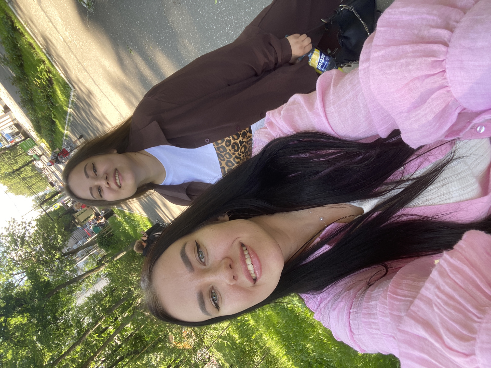 Two women are taking a selfie outdoors, likely on a sunny day. The woman in the foreground on the left has long dark hair, green eyes, and is wearing a pink ruffled shirt. The woman behind her on the right has long brown hair and is wearing a brown open jacket over a white top with a leopard print design. They are positioned in front of a tree-lined path, suggesting a park or a tree-lined street. In the background, other people are visible walking or sitting, and there is a fence and some buildings. A bottle of "Active" brand water is visible in the lower right corner of the image. While the exact location in Perm, Russia cannot be definitively determined from the image alone, the general environment suggests an urban park or public walkway.