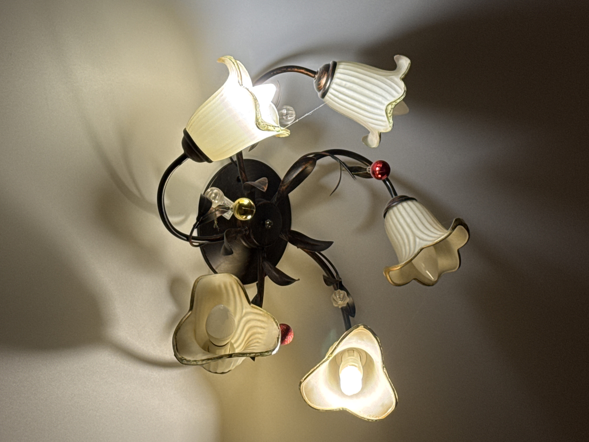 A five-bulb, flower-shaped wall sconce is mounted on a wall. The bulbs are illuminated, casting shadows. The color of the wall transitions from light to dark. This wall fixture cannot be placed in the city of Izhevsk, Russia as no geographic context can be determined from the image.