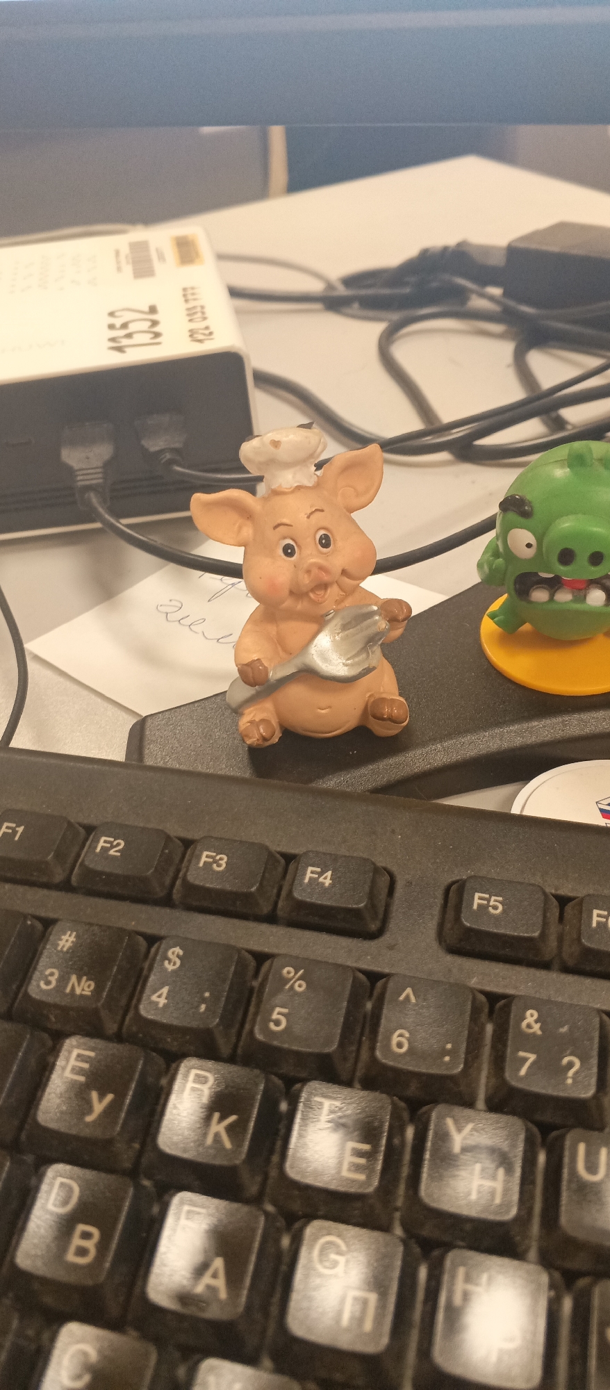 This media file features a close-up, top-down view of a desk setting. The primary subjects are two small figurines: a cartoonish pig dressed as a chef holding a fork and a green Angry Bird character. They are positioned on top of a black computer keyboard.
The scene appears to be indoors, likely an office or workspace, given the presence of the keyboard, computer cables, and a white electronic device with text and a barcode. The lighting suggests an indoor environment, possibly during daytime, but no specific time of day can be determined.
Notable details include the markings on the keyboard, with visible keys labeled "F1" through "F6", as well as various alphanumeric and symbolic keys. Some of these keys also show Cyrillic characters. The white electronic device in the background has the visible text "1352" and "122 035 777". There's also a small piece of paper with handwritten text, partially obscured, which appears to be "глим". The figurines add a playful element to the otherwise utilitarian desk setup. The focus is on the figurines and the keyboard, with the background items slightly out of focus. There are no people visible in the image, and no specific activity or event is clearly taking place beyond the static display of objects.