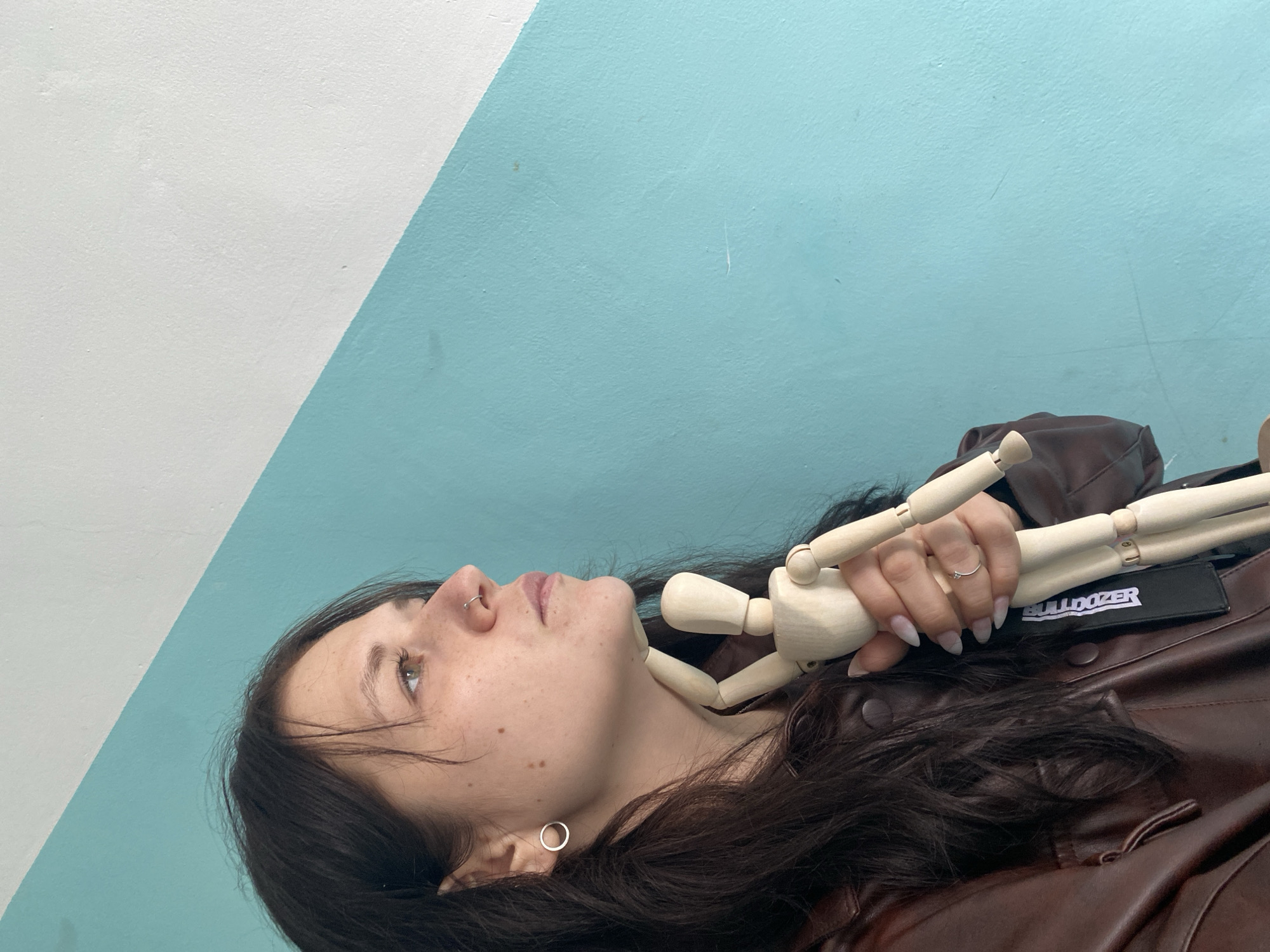 The media file presents a close-up portrait of a young woman against a diagonally split wall, featuring white on the upper left and light blue on the lower right. The woman, with long, dark hair and light-colored eyes, is seen from the chest up, appearing to be lying down or leaning back as she gazes upwards and slightly to her left. Her face shows freckles, and she has a silver hoop nose piercing and a small, round earring in her left ear.
She is wearing a brown leather jacket, on which a black tag with the white text "BULLDOZER" is visible. Her right hand, featuring long, manicured nails with a subtle ombre gradient and a delicate silver ring, holds a small, wooden artist's mannequin. The mannequin rests across her chest and neck area. The wall texture appears slightly coarse. The lighting is soft and even, casting no prominent shadows. The overall mood is serene and contemplative. This moment was captured in Chaikovsky, Russia.