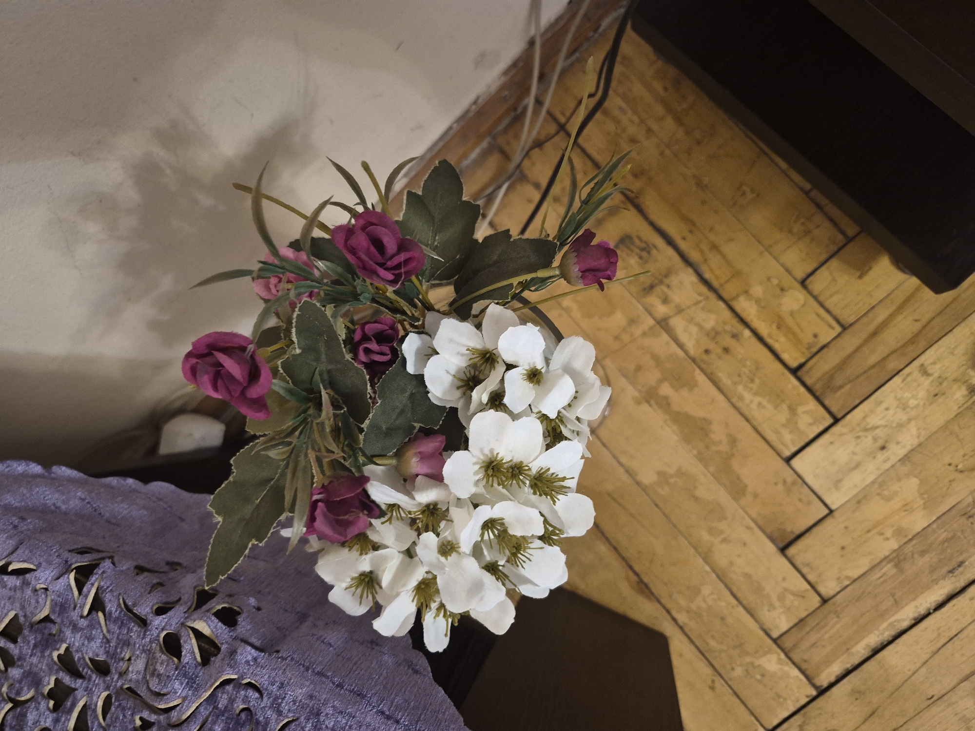 A bouquet of artificial flowers, featuring white blossoms with yellow centers and deep pink roses, is placed in a vase on a wooden floor. The floorboards are laid in a herringbone pattern. To the left of the bouquet, a portion of a purple textile with an ornate, cut-out design is visible, likely a tablecloth or decorative runner. The background consists of a light-colored wall with subtle shadows. The scene is indoors, possibly in a residential setting in Yerevan, Armenia.