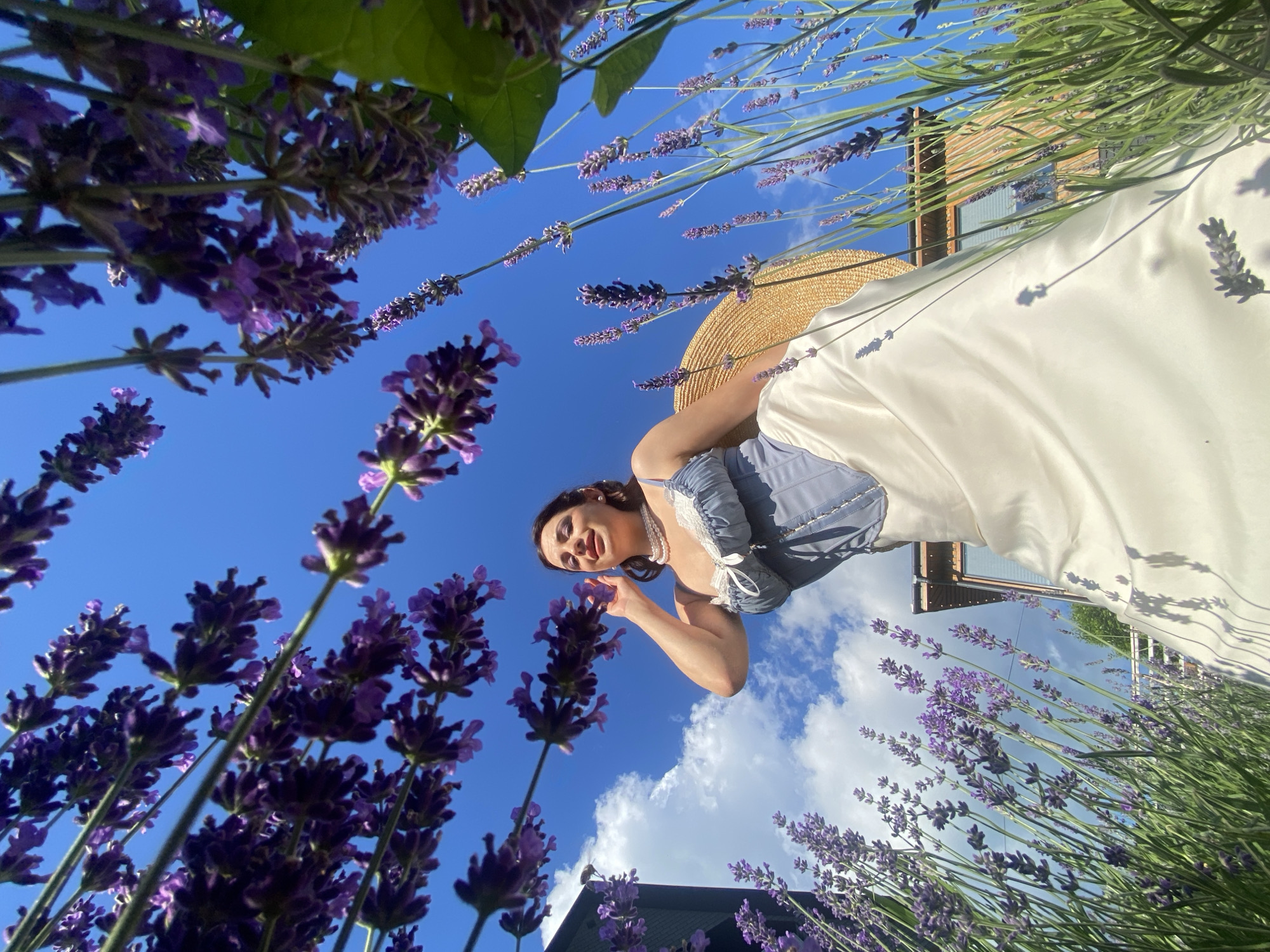A low-angle shot captures a woman in a blue corset and a long white skirt, standing in a lavender field under a bright blue sky. The lavender stalks, with their vibrant purple blooms, frame the woman and extend upwards. She is wearing a pearl necklace and holds a straw hat. In the background, a modern building with wooden accents is visible. The image is taken in Verkhneye Uchdere, Russia.