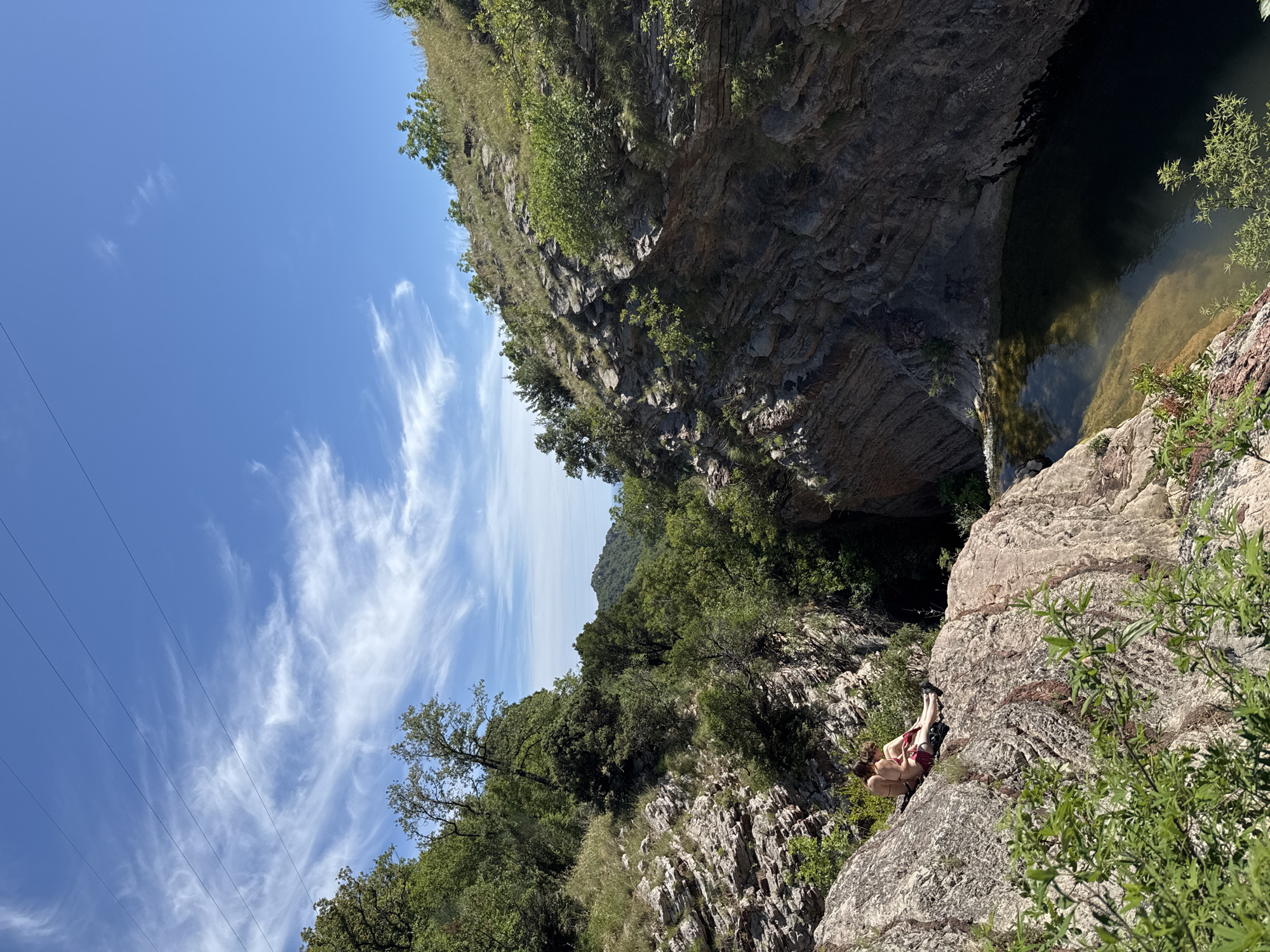 Two individuals are seated on a rocky ledge overlooking a natural pool of water surrounded by steep, forested cliffs. The individuals are casually dressed, with one wearing a bikini and the other in swim trunks. They are positioned close together, facing away from the camera and towards the expansive natural landscape. The environment is characterized by rugged terrain, lush green vegetation, and a clear blue sky with wispy white clouds. In the distance, mountains can be seen, and overhead power lines are faintly visible against the sky. The scene appears to be in a remote, outdoor location, potentially a natural park or a less-developed area of Rafailovići, Montenegro.
