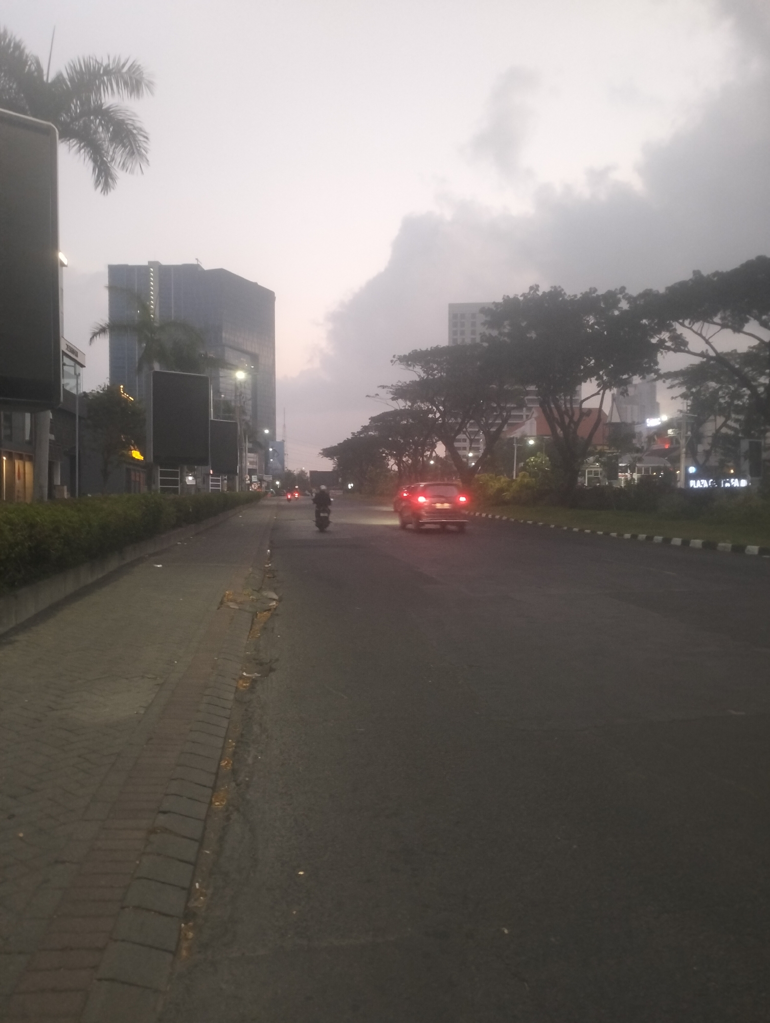 A street scene at dusk in Surabaya, Indonesia, features a road with a motorcycle and a car driving away from the viewer. On the left side of the road, a brick pathway is lined with a low, manicured hedge. Behind the hedge, buildings rise, including a modern, tall glass skyscraper with numerous illuminated windows and a palm tree in front of it. Across the street, on the right, are more trees and a building with a sign that reads "PLAZA." The sky is overcast with thick, grey clouds, and the overall lighting suggests it is either early morning or late evening. The road itself is asphalt, with a distinct curb and a black and white striped border marking the edge. There are some pieces of litter visible on the pathway and road. The car's brake lights are illuminated, and the motorcycle rider is wearing dark clothing.