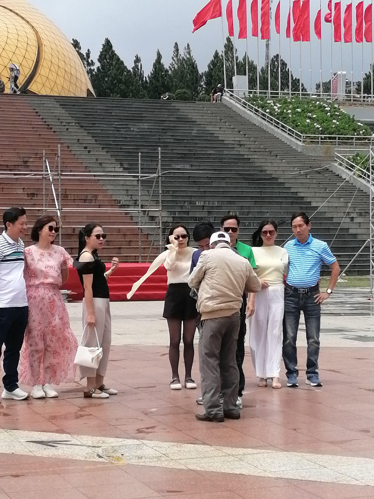 The image depicts a group of people standing on a paved area in front of a large set of concrete stairs. To the left, a golden dome structure is partially visible. In the background, a line of red flags is flying. Several individuals are dressed casually, with some wearing sunglasses. The weather appears overcast. This scene is located at the Quang Trung Flower Park in Da Lat, Vietnam, specifically in the area leading up to the park's main amphitheater or viewing platform. The group appears to be tourists posing for a photograph.