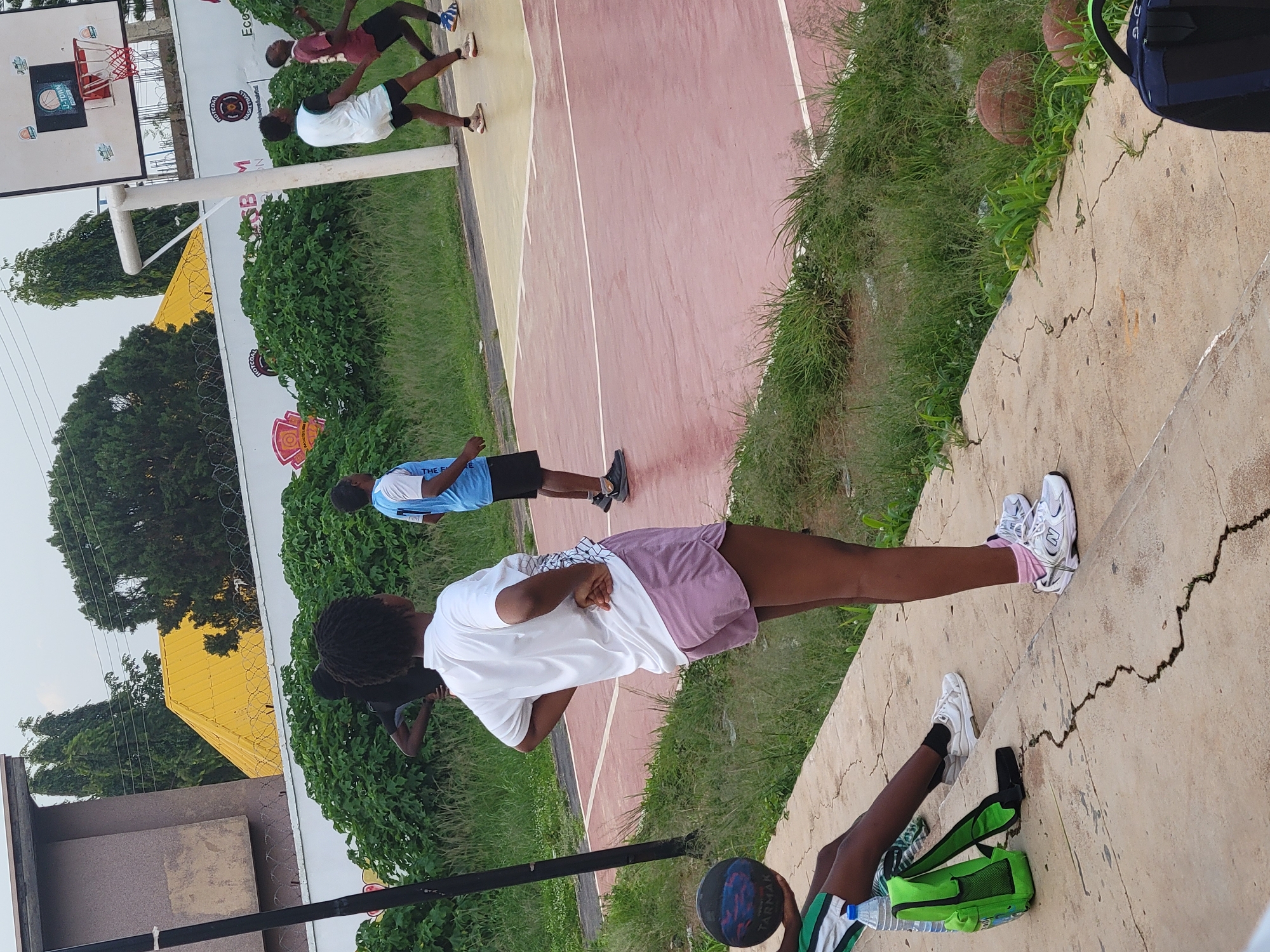 The image shows a group of young people on an outdoor basketball court. There are at least five individuals visible, most of whom are wearing athletic clothing. One person in the foreground is wearing a white t-shirt and purple shorts, with white sneakers and pink socks. Another person is wearing a light blue jersey and black shorts. Further in the background, two individuals in maroon and white shirts are also on the court. A basketball is visible in the lower right portion of the image, near a green bag and a water bottle. The court itself is made of reddish and yellowish material with white lines. The setting appears to be daytime, with overcast skies, and there is ample green grass and trees surrounding the court. The people seem to be engaged in some sort of physical activity, possibly a training session or practice. There are basketball hoops and backboards visible in the background. The overall mood seems to be active and casual. The text "TARMAK" is visible on the basketball.