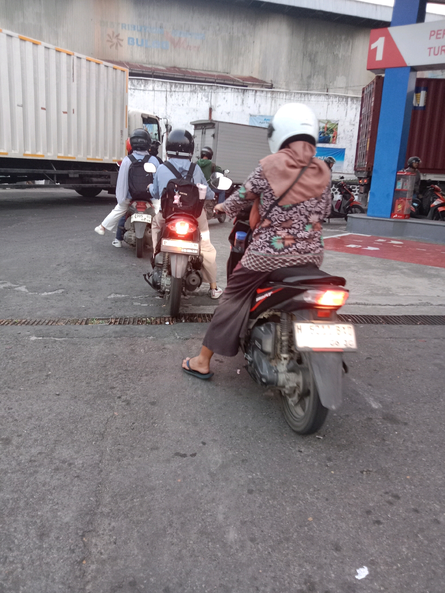 The image depicts a street scene in Semarang, Indonesia, likely a busy area near a gas station, as indicated by a sign with the number "1" and the word "PERTAMINA" (a state-owned oil and gas company in Indonesia) visible in the background. Several people are on motorcycles, waiting in what appears to be a queue. The perspective is from behind the motorcyclists, focusing on their backs and the rear of their vehicles.
In the foreground, a person on a motorcycle wearing a patterned brown and pink top and dark pants is prominent. They are wearing a white helmet. The motorcycle's rear brake light is illuminated. A license plate is visible, reading "H 5211 SBD" and a date below it. Another motorcycle behind them has a license plate reading "2305 RU" with a date. The rider of this motorcycle wears a light blue long-sleeved shirt and a black backpack. Further back, another motorcyclist in a similar light blue shirt is visible.
The environment appears to be a paved road with some debris. In the background, there is a large white truck with a ribbed trailer, a building with a corrugated metal exterior, and what seems to be a gas pump structure. The overall impression is a moment captured in the daily life of traffic in Semarang.