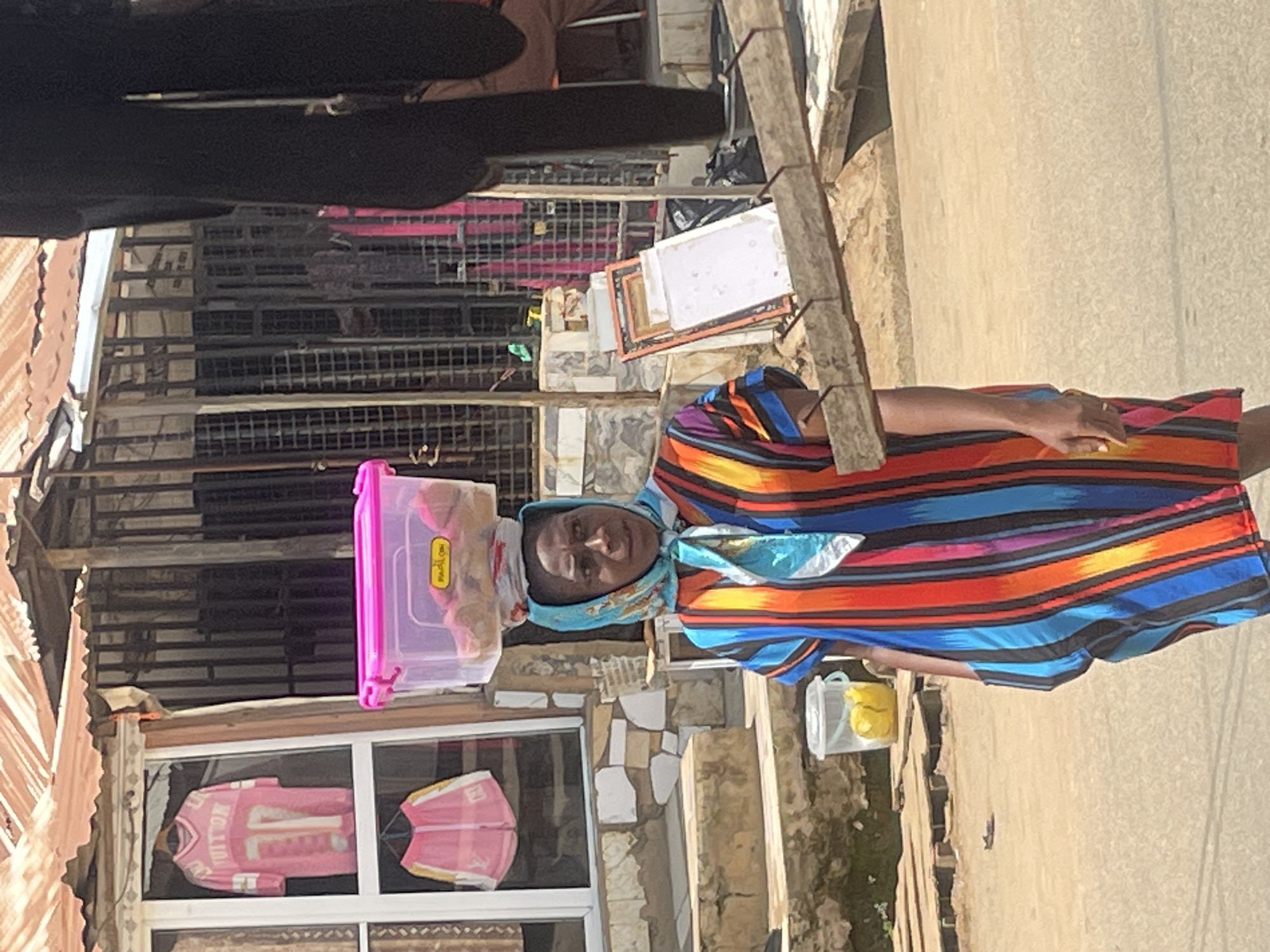 A Nigerian woman is captured in a medium shot, walking outdoors in what appears to be a market or street scene in Abuja, Nigeria. She is wearing a vibrant, vertically striped dress in shades of blue, orange, red, and purple, and a light blue and white patterned headscarf tied around her neck and head. The woman is carrying a pink plastic container filled with food items balanced on her head. She is looking slightly to her right, with a neutral expression.
The background consists of a building with a corrugated metal roof and an open storefront displaying two pink garments. To the right of the woman, a dark, textured surface suggests the presence of another person or structure. In the foreground, a wooden plank with nails is laid across two metal poles, partially obscuring the ground and a small plastic bucket containing yellow objects. The ground is paved with a rough, sandy texture. The overall lighting suggests a daytime scene with bright sunlight.