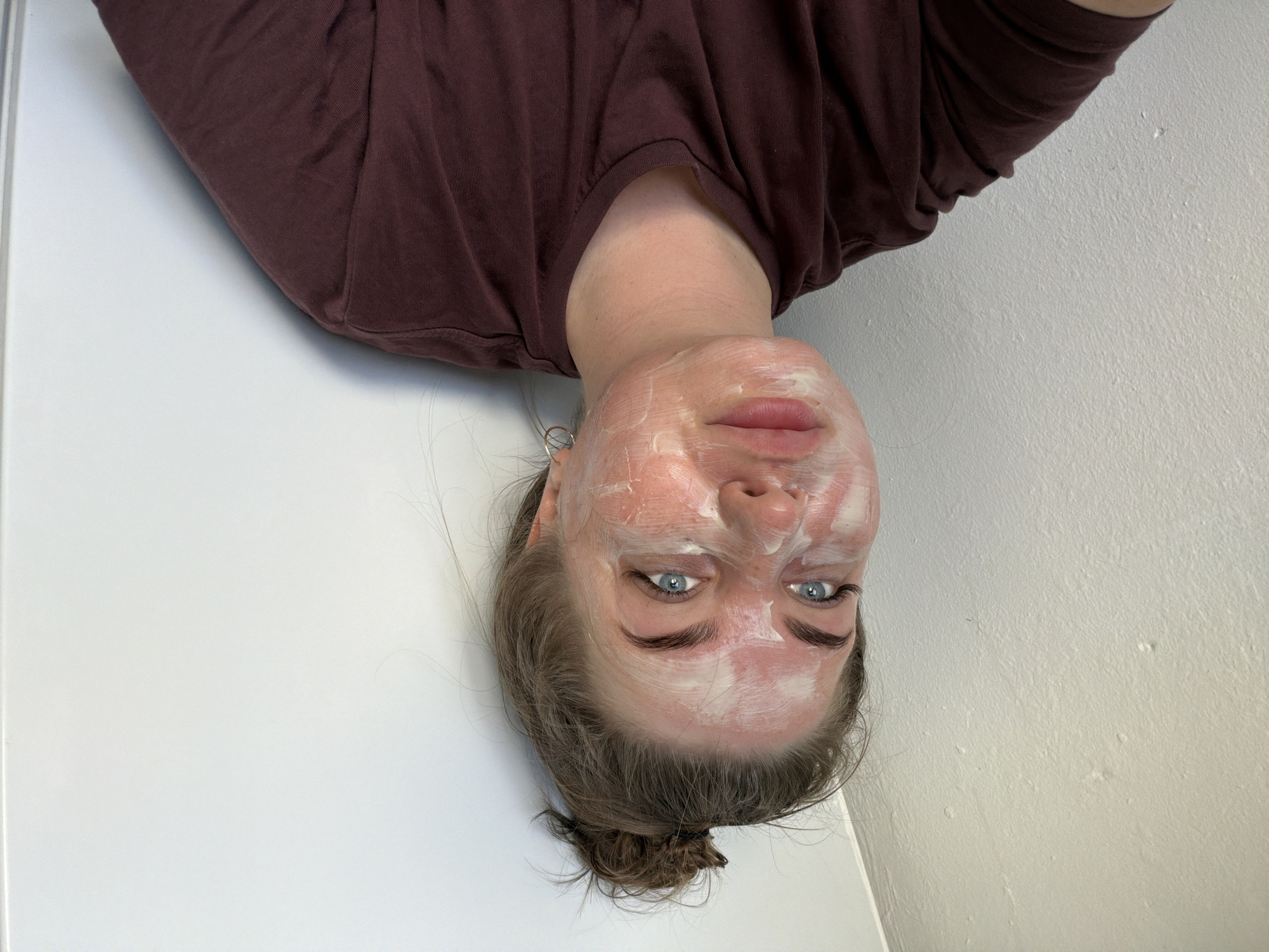 A woman wearing a dark burgundy t-shirt lies on a white surface, presumably indoors, as evidenced by the adjacent white wall. Her hair is pulled back into a bun. She has a white cream facial mask on her face, covering her forehead, cheeks, nose, and chin. She has light blue eyes. Her location in the city of Chaikovsky, Russia, cannot be determined from the given image.