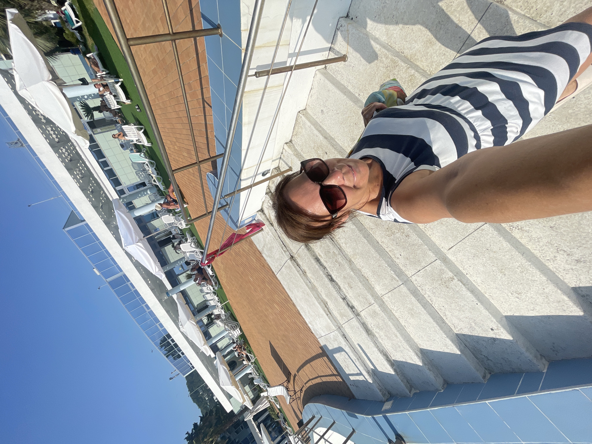 A woman with dark hair and sunglasses is taking a selfie on a sunny day in Sochi, Russia, likely at a resort or hotel with a pool area. She is wearing a striped dress and holding a bag. In the background, there are lounge chairs, umbrellas, and a building with glass windows, suggesting a recreational area. The stairs she is standing on appear to be made of concrete and have blue tiled accents around the pool. The sky is clear and blue.