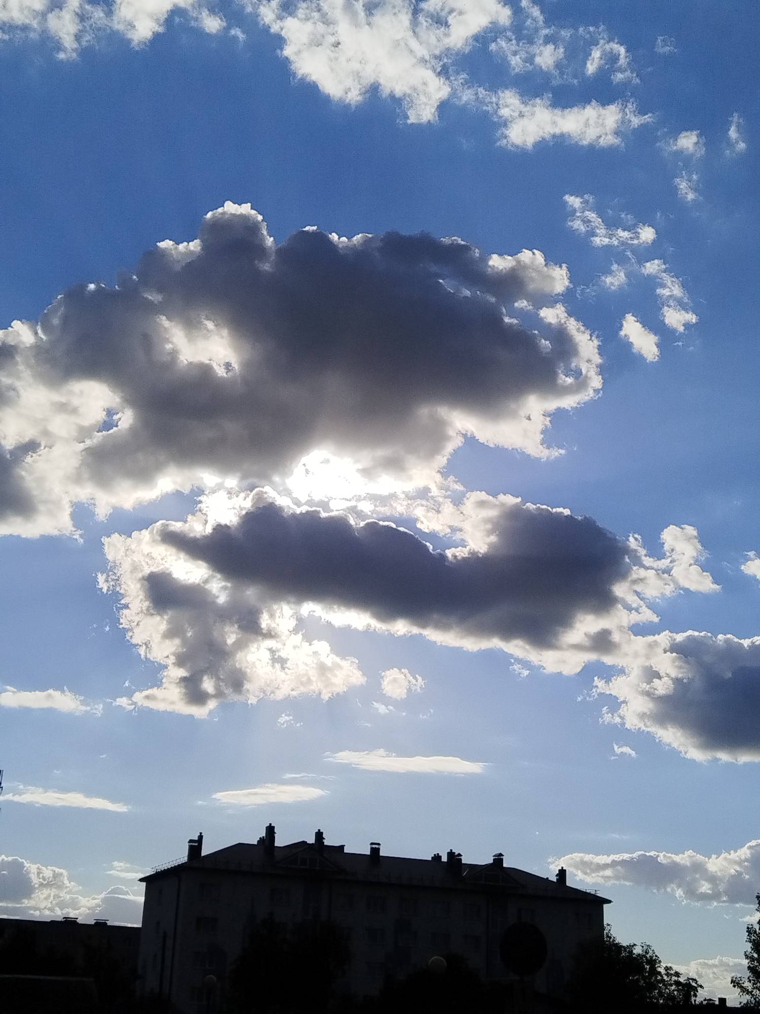 This is a vertical, eye-level, close-up shot of a cloudy blue sky with a building silhouette in the foreground, likely taken in the city of Brest, Belarus. The sky is a vibrant blue, with scattered white and grey clouds of various sizes. Some of the larger grey clouds are dramatic, with sunlight piercing through them, creating a sense of depth and brilliance. The sun itself is obscured by the clouds, but its light is clearly visible.
In the lower third of the image, a long, dark silhouette of a multi-story building stretches across the frame. The building appears to be of typical Soviet-era architecture, with a flat roof and visible chimneys. The upper edges of trees are also visible, silhouetted against the sky. The overall impression is a dramatic sky scene with a subdued, man-made element in the foreground. The lighting suggests it is daytime, possibly late afternoon or early evening, given the contrast between the illuminated clouds and the shadowed building.