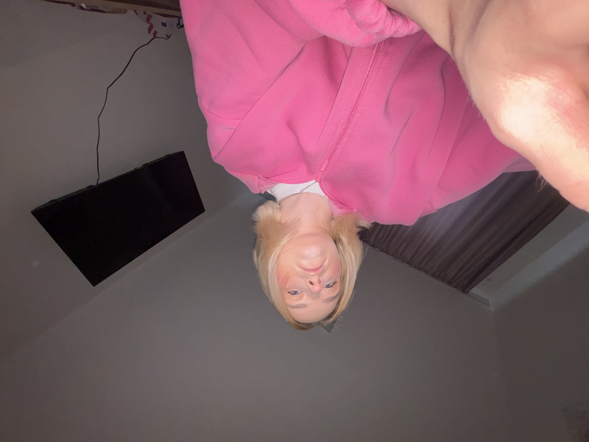 A photo shows a person wearing a pink zippered sweatshirt, with a television and electrical wire visible on the wall. The person has blonde hair and fair skin. The room appears to be a bedroom. The specific location in Vsevolozhsk, Russia, cannot be determined from the image.