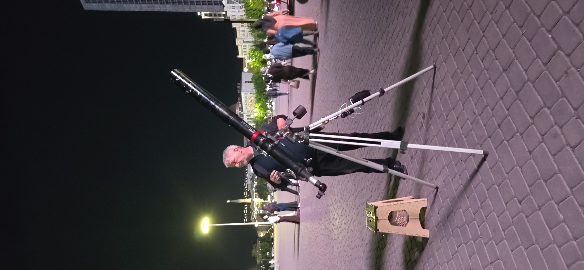 The scene depicts a man standing next to a large telescope on a tripod, likely set up for public viewing. He appears to be adjusting or operating the telescope. A small stool is positioned near the telescope's tripod. Several people are blurred in the background, suggesting movement and a public space. The background also includes street lights and building structures, indicating an urban setting at night. Based on these observations, it is inferred that the location is the city of Grozny, Russia. The photo was taken at nighttime outdoors.