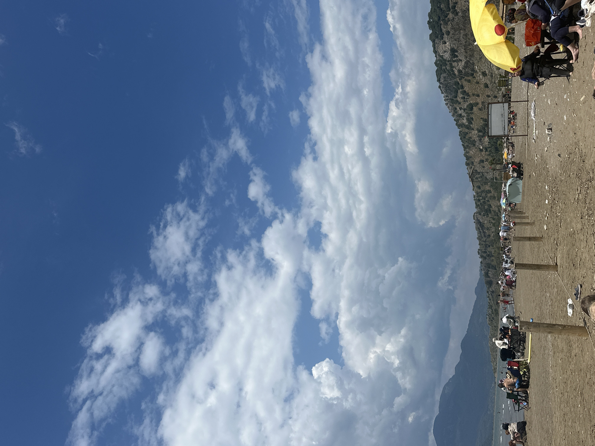 The image depicts a beach scene in Ortaca, Turkey. A mountain range borders the beach. The sky is partly cloudy, with a section of deep blue. The beach is populated with people, many under umbrellas. There is an information board present on the beach. Wooden posts line the beach. People are sitting in chairs along the beach.