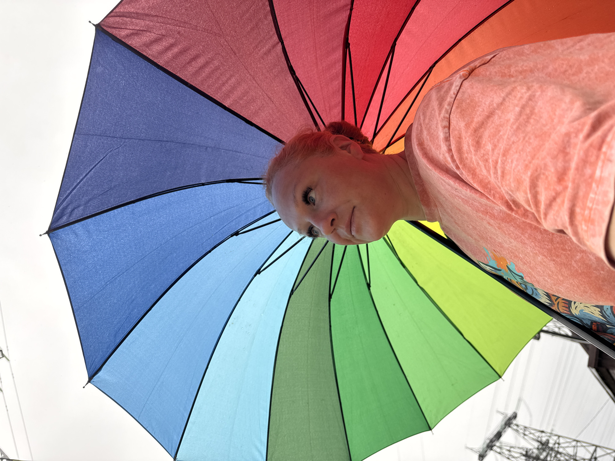 A low-angle shot of a person standing under a large, multicolored umbrella. The umbrella is comprised of red, orange, yellow, green, blue, and indigo segments. The person, presumably a woman, has light skin and reddish hair. She is wearing a salmon-colored shirt. The background reveals a cloudy sky and power lines, possibly suggesting an urban or suburban setting in Chaikovsky, Russia.