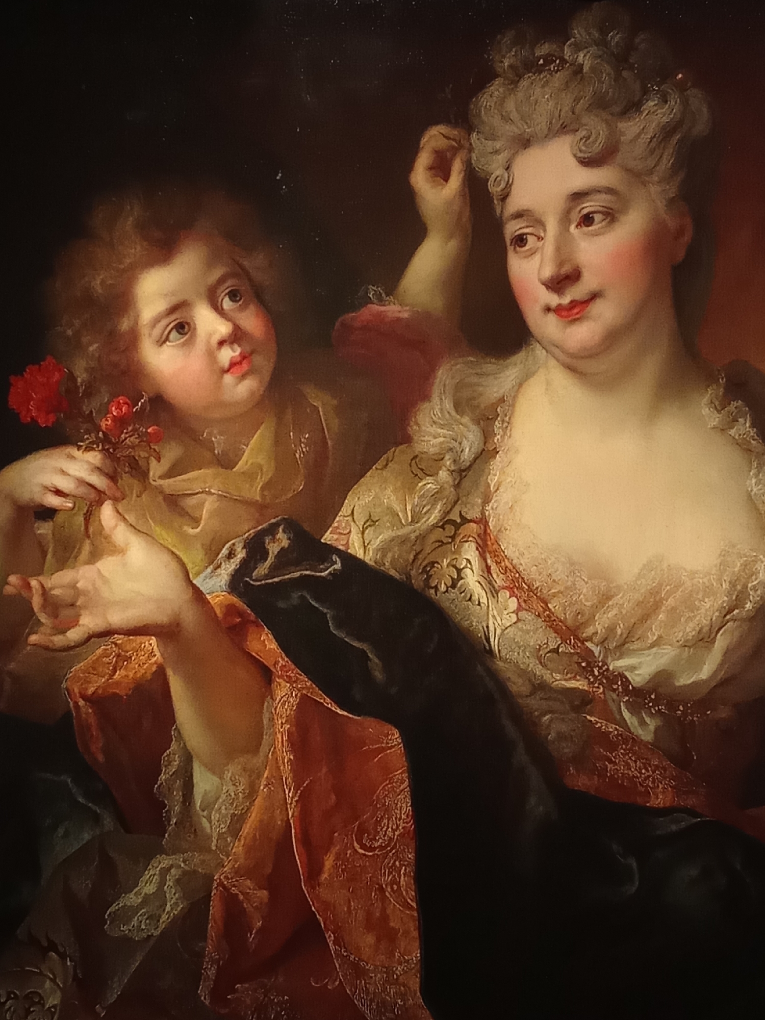 This is a painting depicting a woman and a child. The woman, on the right, is older with powdered, upswept hair adorned with pearls. She has rosy cheeks and is wearing a low-cut, lacy dress with gold and floral embroidery. Her left hand is raised, touching her hair, while her right hand is extended towards the child. The child, on the left, appears young, with rosy cheeks and wide eyes looking upwards. They are holding a small bouquet of red flowers in their left hand and have their right hand outstretched, palm up, in a gesture of offering or reaching. The child is dressed in a yellow garment. The background is dark, with warm tones suggesting a draped fabric or interior setting.
The painting is located in the Museo Nacional de Bellas Artes in Buenos Aires, Argentina.
