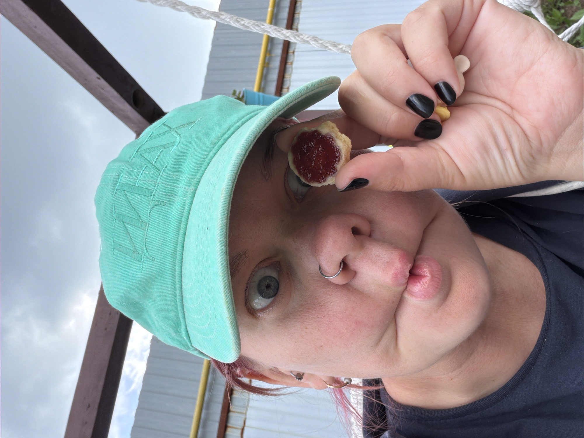 A close-up, outdoor, selfie-style photograph shows a person wearing a teal baseball cap and a dark t-shirt. The person has pink hair visible beneath the cap, a nose ring, and an earring in their left ear. Their eyes are wide open, and their lips are pursed. The person is holding a small, round cookie topped with red jam between their thumb and index finger, bringing it close to their face. Additional small, light-colored objects, possibly candies or pills, are held between their fingers. In the background, a wooden structure, a corrugated metal fence, and a rope from what appears to be a swing set are visible. The sky above is overcast. The image was taken in Chaikovsky, Russia, indicated by the presence of a yellow gas pipe, a common feature in Russian residential areas.