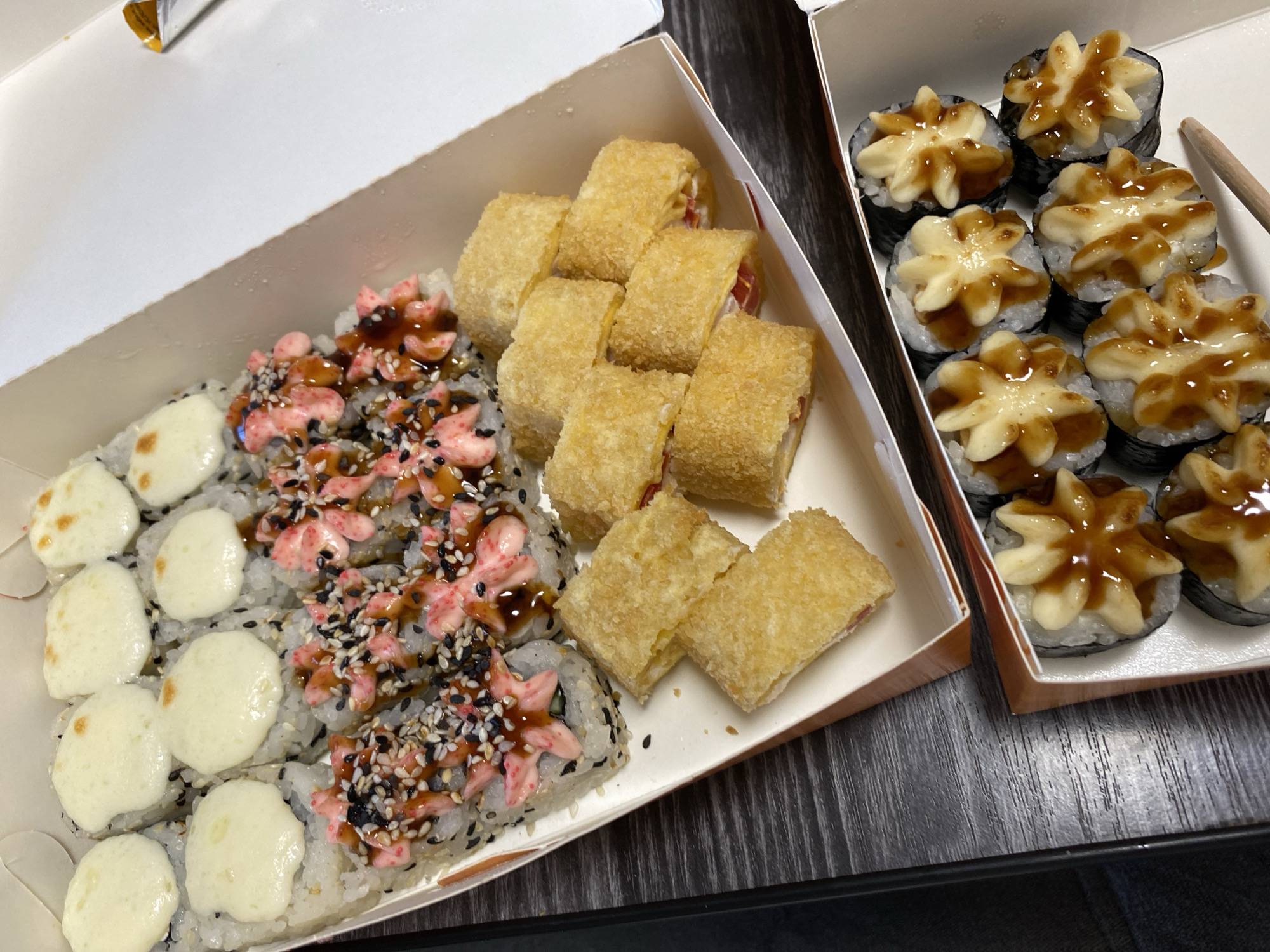 Two white rectangular takeout containers filled with different types of sushi are positioned on a dark, possibly wooden, surface. The left container holds three distinct sushi variations: pieces topped with melted cheese, others with pink sauce and black sesame seeds, and tempura-style sushi with a golden-brown, fried coating. The right container presents sushi rolls adorned with cream-colored, flower-shaped toppings and a brown drizzle. A wooden utensil rests beside the sushi in the right container. The setting appears to be indoors. The image does not offer enough detail to identify a specific location within a city.