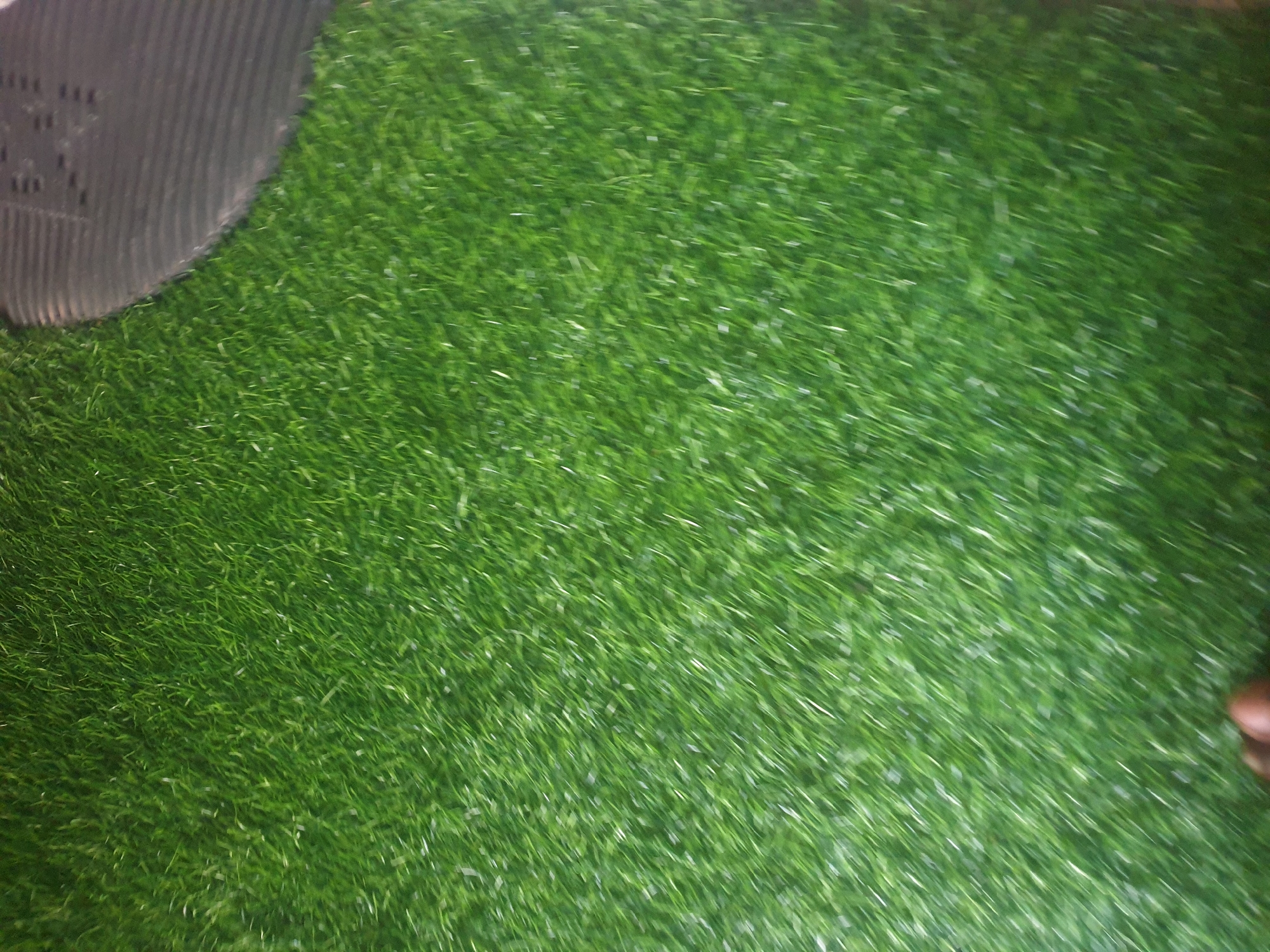 The image shows a close-up, overhead view of artificial green grass. In the upper right corner, the edge of a dark, textured object, possibly a shoe or mat, is visible. In the bottom center, a portion of a person's foot, with brown skin, is also visible. The lighting is dim, and the image appears to have been taken in low-light conditions. There is no indication of a specific location within a city.