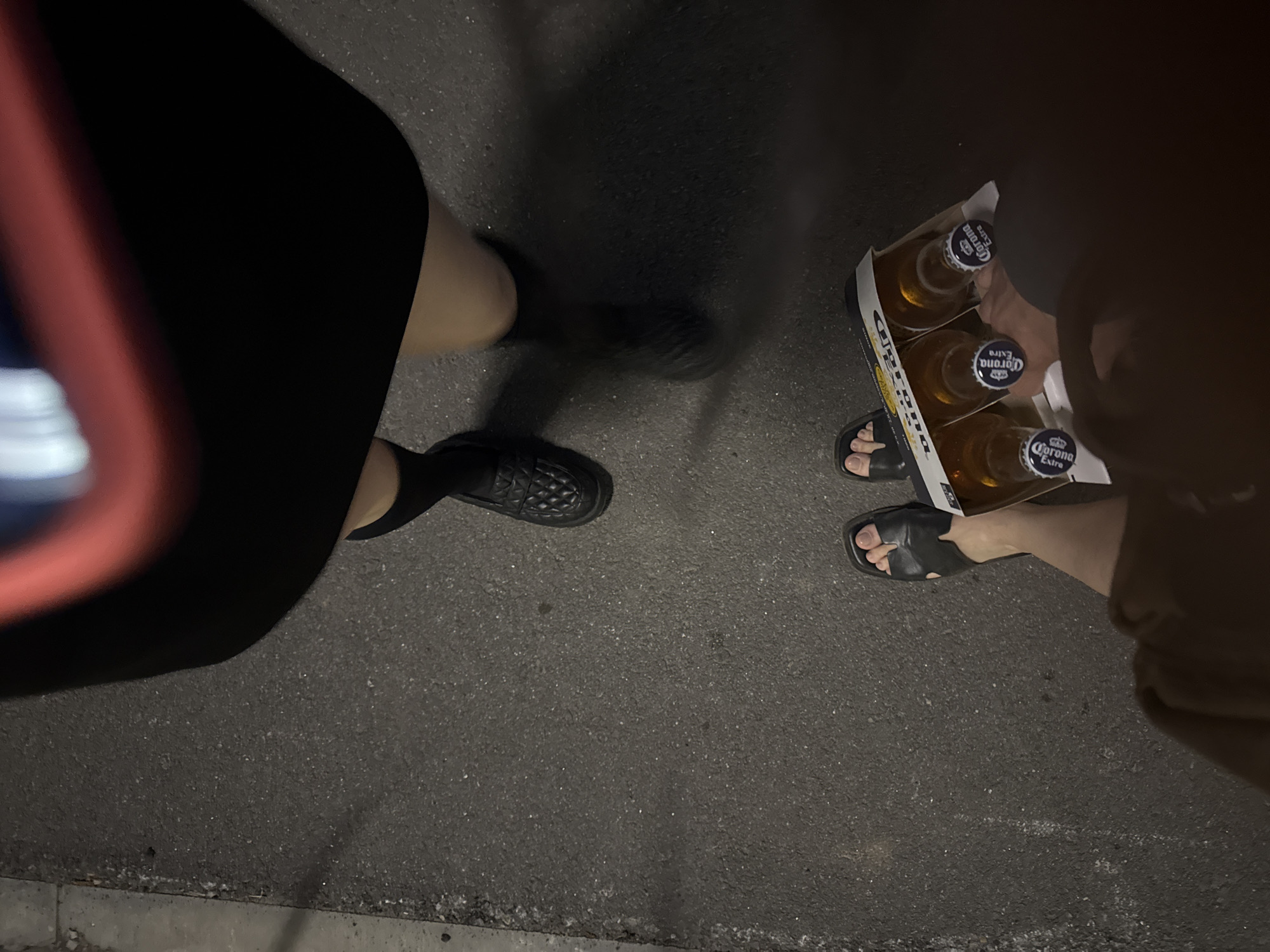 A low-angle, overhead shot shows two people standing on a dark asphalt surface, likely a street. One person on the left is wearing a black skirt and black shoes. The other person on the right is holding a six-pack of Corona Extra beer bottles and is wearing sandals. The background is indistinct, but the asphalt surface suggests an urban environment. Location is unknown.