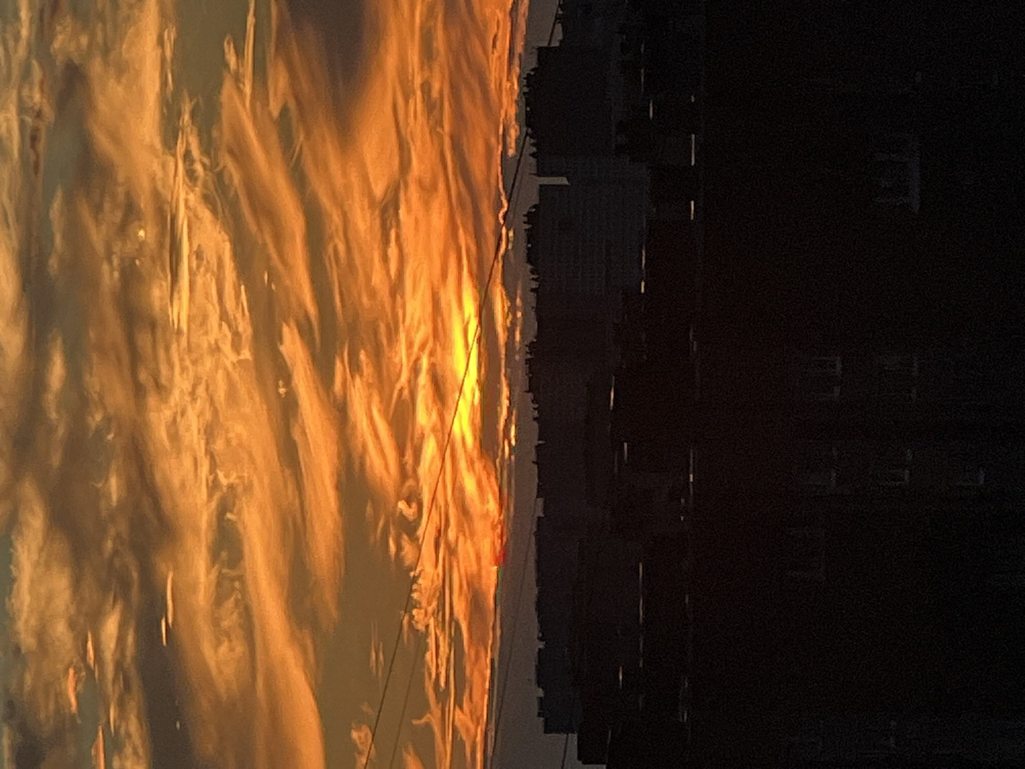 The media file captures a dramatic sunset over a city skyline. The left side of the image is dominated by an intense orange and yellow sky, filled with wispy, illuminated clouds that create a fiery, textured effect. A thin, red sliver of the setting sun is visible just above the horizon line, peeking through the clouds.
To the right of the vibrant sky, the dark silhouettes of buildings rise against the colorful backdrop. The buildings appear to be a mix of modern and older structures, with some exhibiting blocky, rectangular forms and others showing a more detailed architectural outline. The sky transitions to complete darkness on the far right, suggesting the fading light and the approaching night.
The overall scene evokes a sense of awe and possibly melancholy, due to the stark contrast between the brilliant, ephemeral beauty of the sunset and the looming, dark urban landscape. The image is oriented vertically, as if capturing a wide panorama with a tilt. The time of day is clearly sunset. No people or specific objects are discernible beyond the general shapes of buildings. There is no visible text in the image. The location is noted as Unknown, Russia.