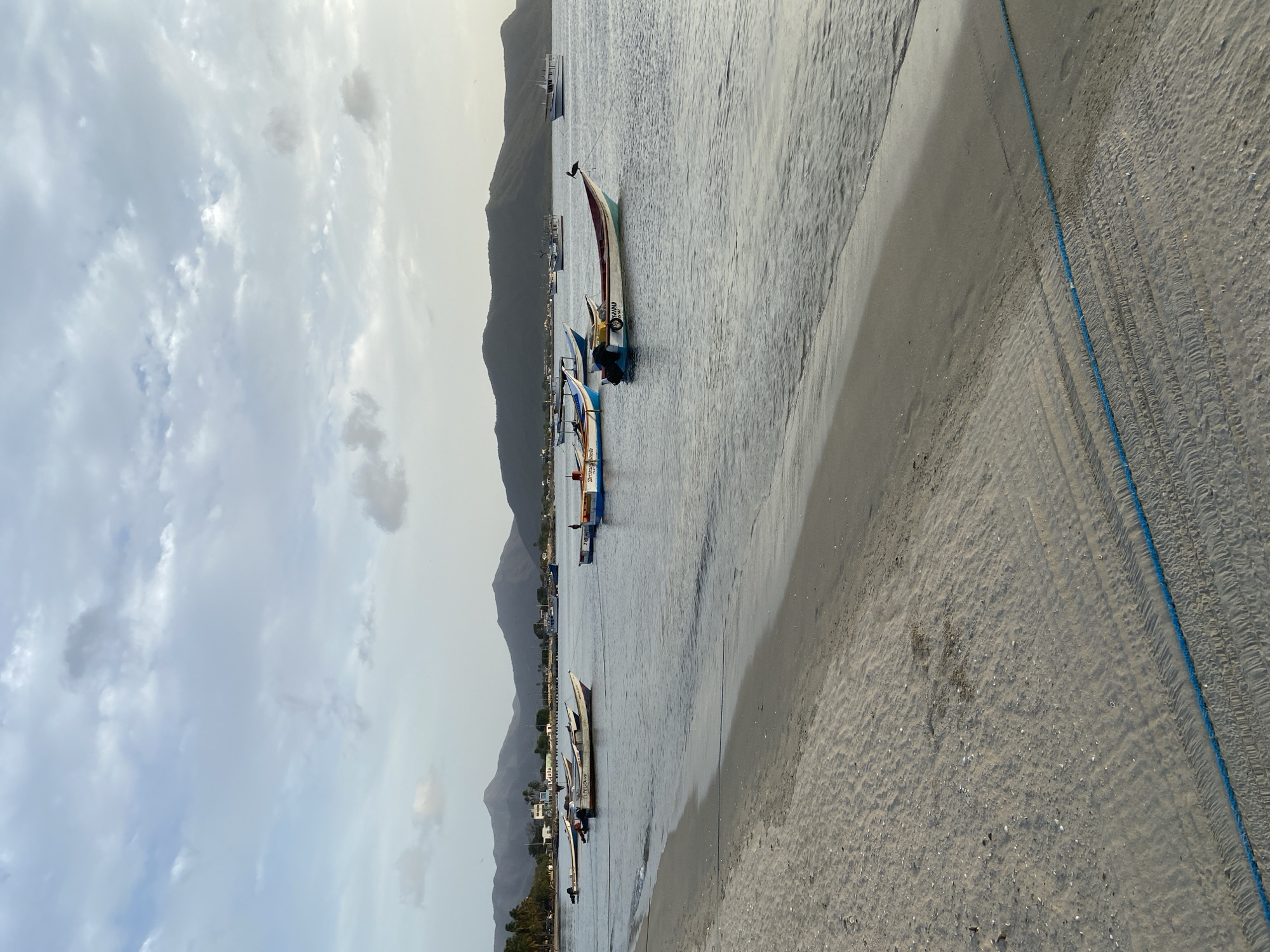 This is a vertical, eye-level shot of a beach scene in Juan Griego, Venezuela. In the foreground, a sandy beach is visible, with a blue rope running horizontally across it. The water is calm and shallow, with gentle waves lapping at the shore. Several small fishing boats are anchored in the water, some with their engines attached. In the background, a chain of mountains rises behind the town, which is sparsely populated with buildings and palm trees. The sky is overcast with clouds, and the overall lighting suggests it is either early morning or late afternoon. A solitary bird is perched on the bow of one of the boats.