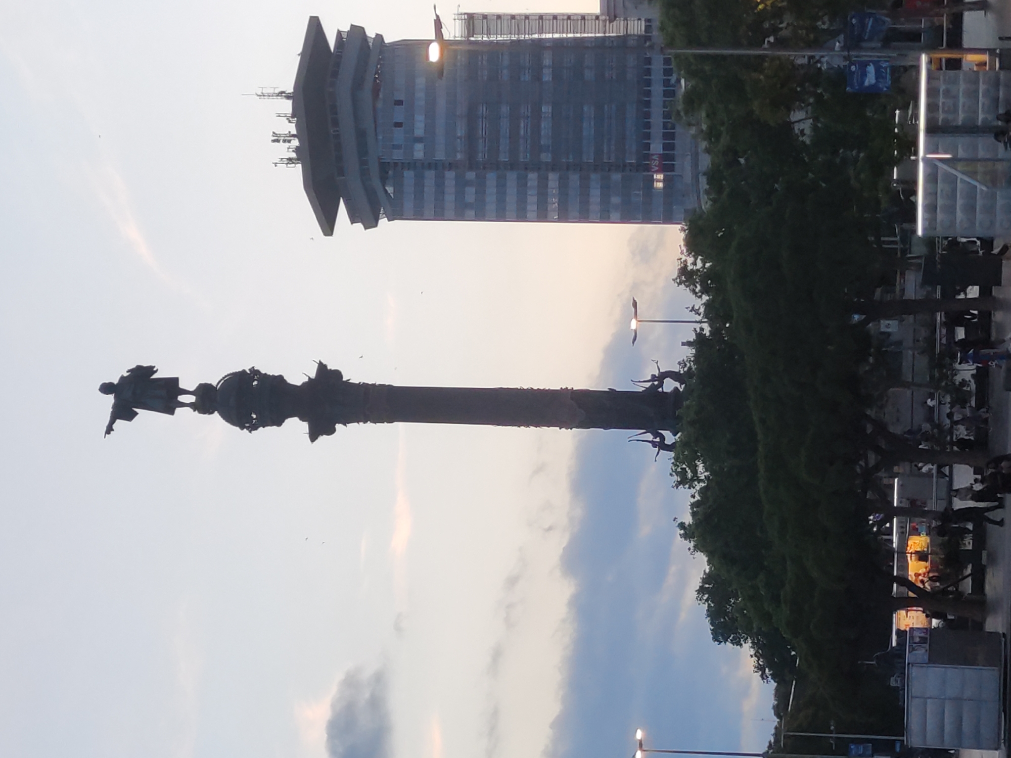 The image depicts a view of the Columbus Monument in Barcelona, Spain, with a modern high-rise building in the background. The monument, a prominent landmark in the city, features a statue of Christopher Columbus pointing towards the sea, standing atop a tall column. The sky is overcast, suggesting it may be late afternoon or early evening. In the foreground, trees and a few people are visible, along with a food cart. The high-rise building is characterized by its glass facade and multiple antennas on its roof. The scene conveys a sense of urban landscape combining historical monument with contemporary architecture. The question incorrectly states the location as Sant Boi de Llobregat, Spain. This monument is located in Barcelona, Spain.