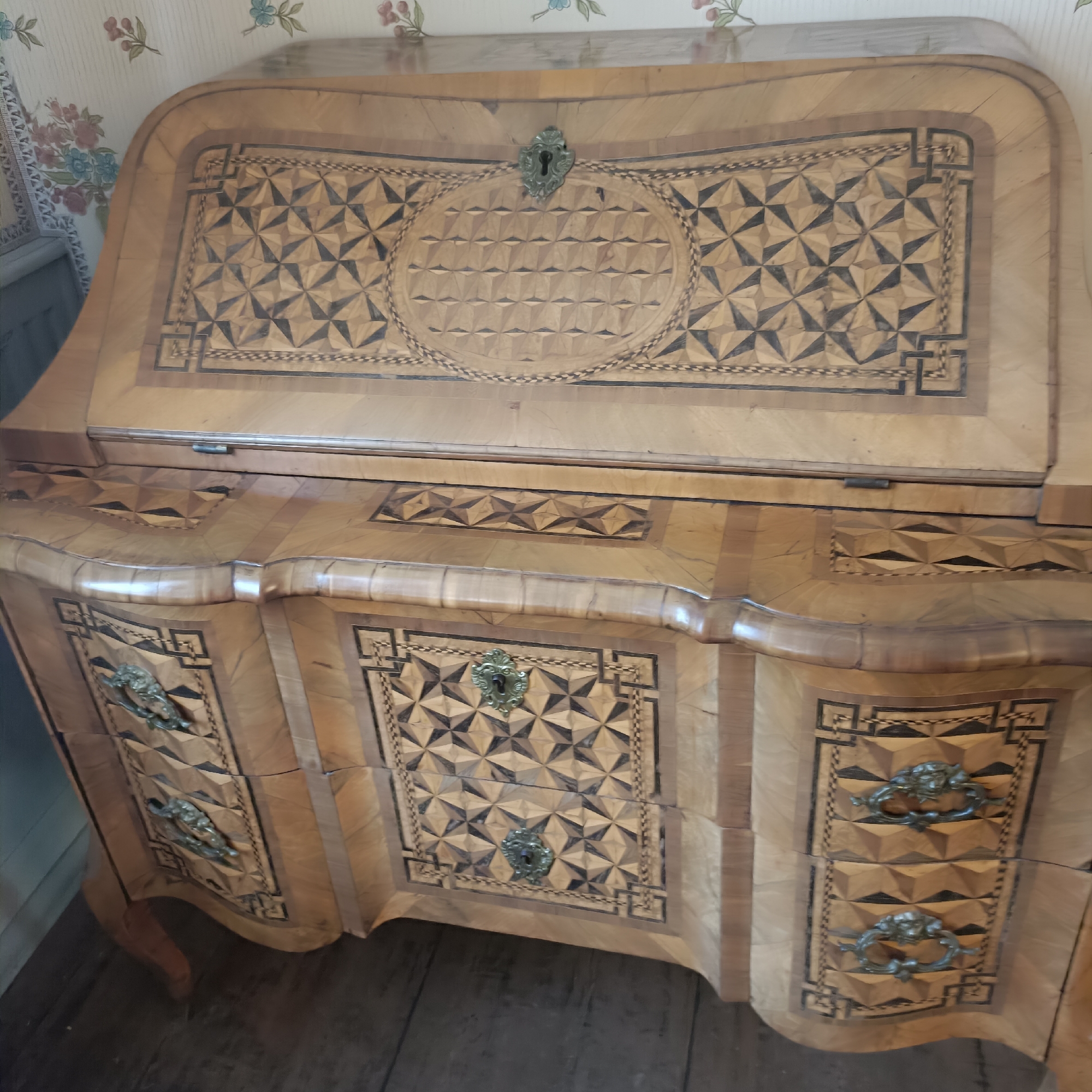 The image shows a close-up view of a wooden antique secretary desk. The desk is ornately decorated with inlaid geometric patterns made of light and dark wood. The top surface of the desk features a large oval inlay with a similar geometric pattern and a brass keyhole. Below the top surface are drawers with decorative brass handles and more inlaid patterns. The desk is situated indoors, with a floral patterned wallpaper visible in the background. The floor appears to be made of wooden planks. There are no people or animals present in the image. The lighting suggests an indoor setting, possibly a museum or an antique shop. The image quality is decent, with good detail on the intricate woodwork of the desk.
This image does not depict a scene in the city of Frankfurt, Germany.
