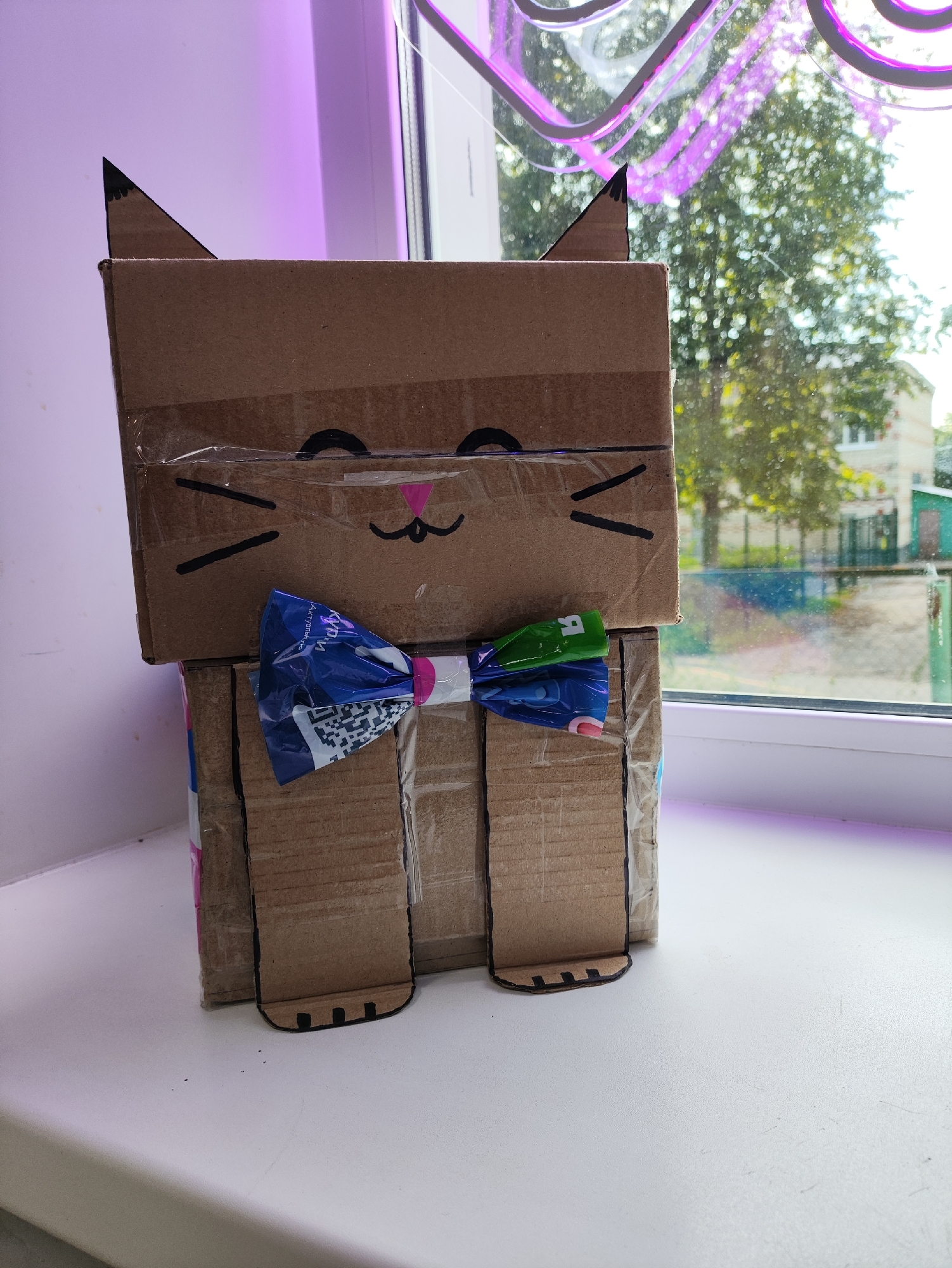 This is a photograph taken indoors, likely in an apartment in Нива, Russia, depicting a cardboard craft project resembling a cat. The craft is positioned on a white windowsill, with a window in the background revealing trees and a building. The cat craft is made from brown cardboard boxes, with black marker details for eyes, nose, and mouth, and black triangles for ears. It also features a blue and green bow tie made of colorful paper or foil. The craft is secured with transparent tape. The ambient light suggests daytime, with a purple light source present, possibly a grow light, casting a glow on the upper left portion of the image. There are no people or animals present in the scene.