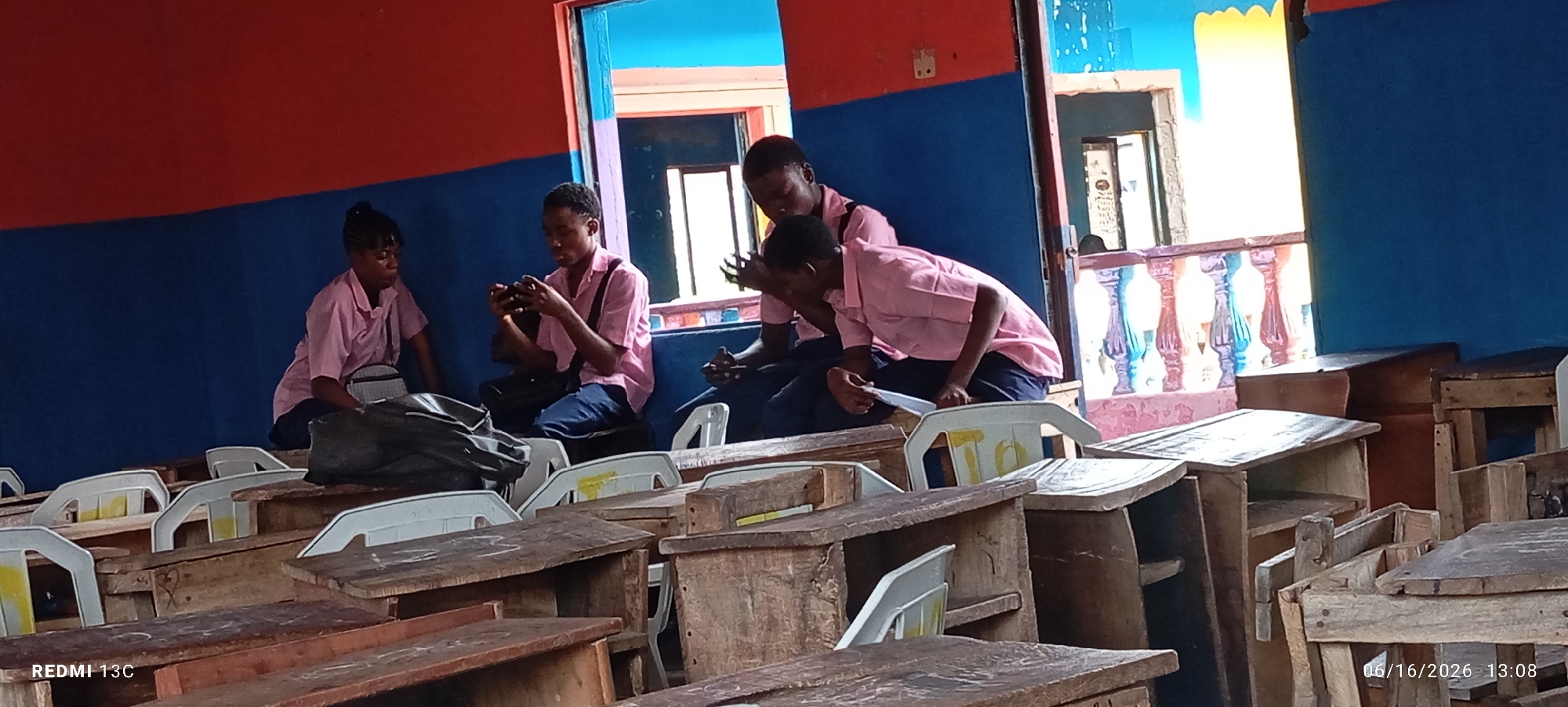 In a classroom setting with a bright red and blue painted wall, four young people, likely students, are seated on chairs in front of wooden desks. Three of them appear to be wearing light pink collared shirts, suggesting a school uniform. The student on the far left is looking down at their lap. The second student from the left is holding a mobile phone and looking at it. The third student from the left is also holding a mobile phone and looking down. The fourth student, on the far right, is looking down at something in their hands, possibly a piece of paper. The classroom is sparsely furnished, with empty white plastic chairs visible between the desks. A doorway with colorful painted trim is visible in the background, revealing an outdoor space with a decorative railing. The image appears to have been taken in Lagos, Nigeria, given the presence of the "REDMI 13C" watermark and the date "06/16/2026 13:08" indicating a future date, possibly a data error or a placeholder, but the device model is commonly used in Nigeria.