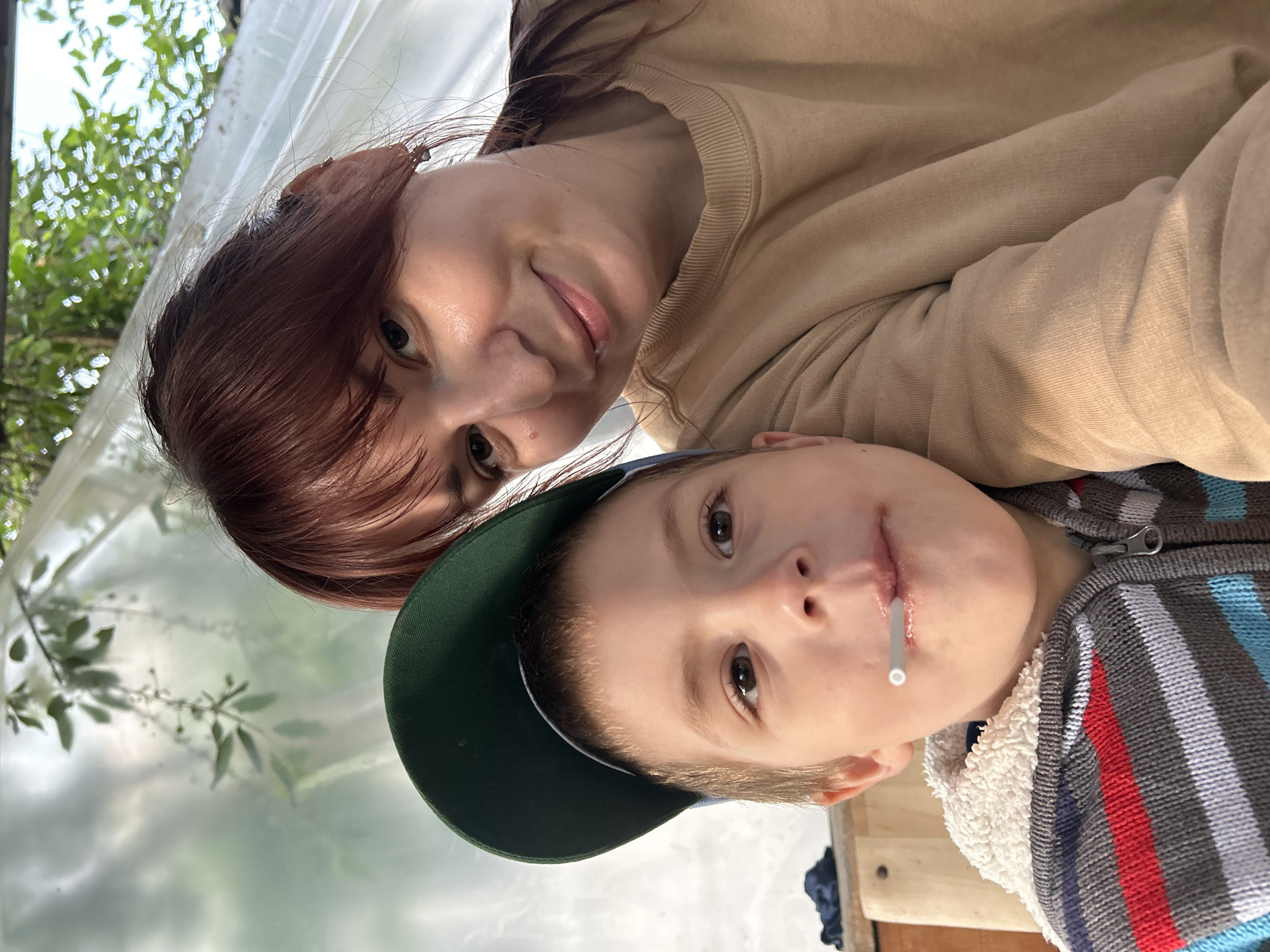 A woman and a child are posing for a selfie. The woman has shoulder-length red-brown hair and is wearing a tan-colored sweatshirt. The child is wearing a green baseball cap and a striped sweater. There is a white object in the child's mouth. The background appears to be inside a greenhouse, with blurry greenery visible through the translucent plastic. The location cannot be determined from the image.