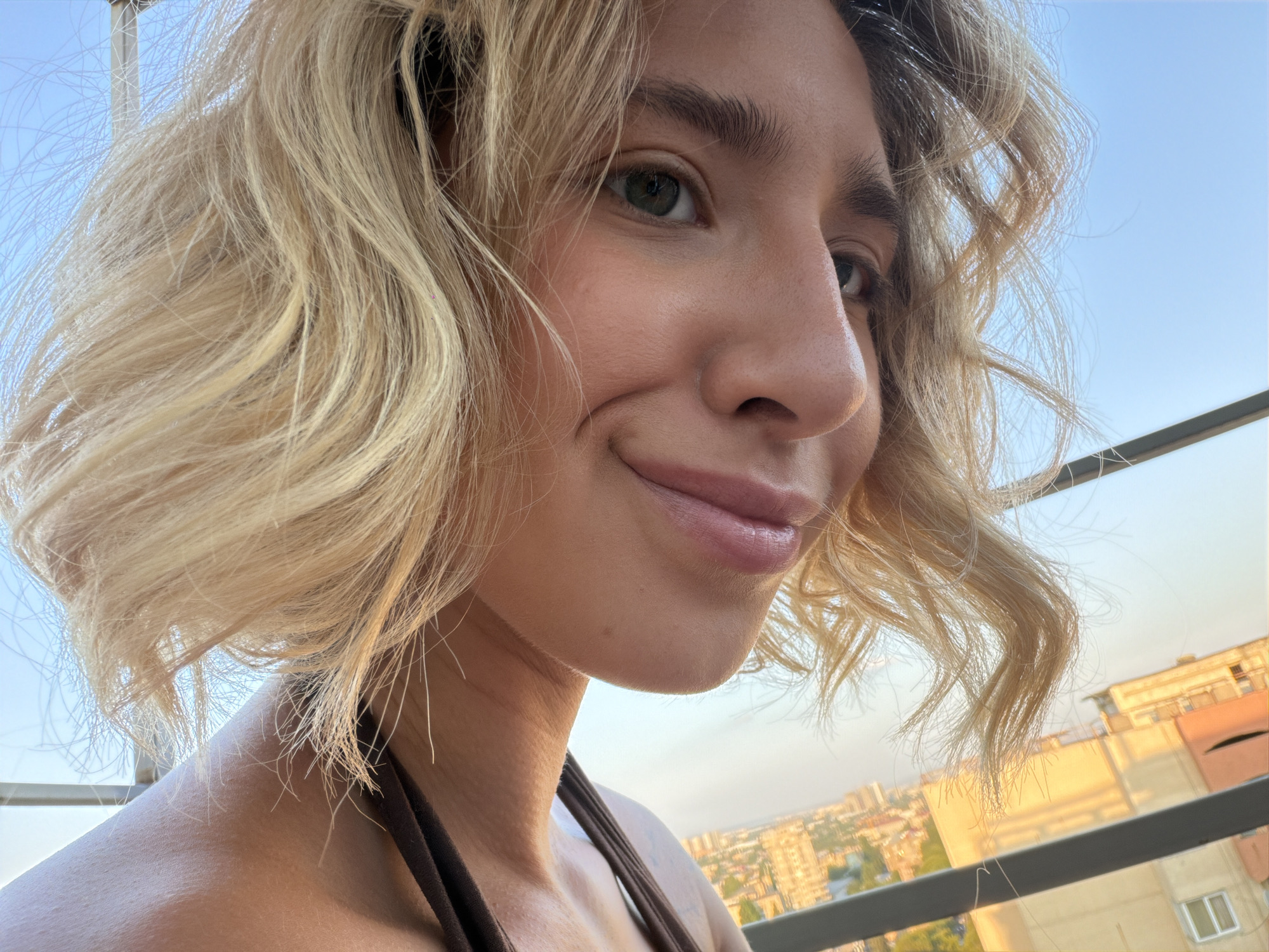 A close-up, profile shot captures a young Caucasian woman with short, blonde, wavy hair against a clear blue sky. She is looking to the right, with her head tilted slightly. Her eyes are green, and she has a faint smile, with her lips slightly parted. Her skin appears smooth and lightly tanned. She is wearing a dark brown top.
The background shows a city skyline, consistent with Tbilisi, Georgia, with buildings visible in the distance. A railing runs across the bottom of the frame, suggesting she is on a balcony or rooftop. The lighting suggests it is late afternoon or early evening, as the sun casts a warm glow.