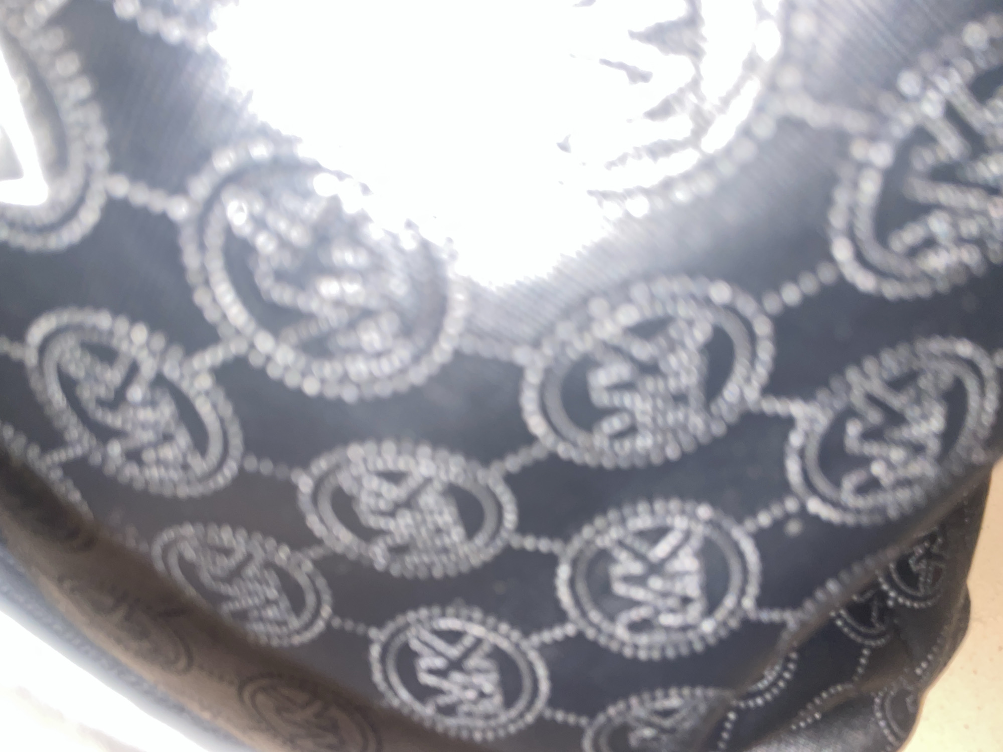 The image displays a close-up, blurry view of a dark fabric with a repeating pattern of interlocking circular shapes. Each circle contains what appears to be a stylized logo or monogram, possibly the letters "MK." The pattern is rendered in a lighter color, creating a contrast against the dark background. The fabric seems to be textured, with the pattern possibly embossed or printed. The lighting in the image is very bright, particularly in the upper half, creating a washed-out effect and making it difficult to discern fine details. The lower portion of the image shows some curvature of the fabric, suggesting it might be part of a bag or a similar item. No people, specific activities, or clear environmental cues are visible due to the focus and lighting. There is no discernible text beyond the potential logo.