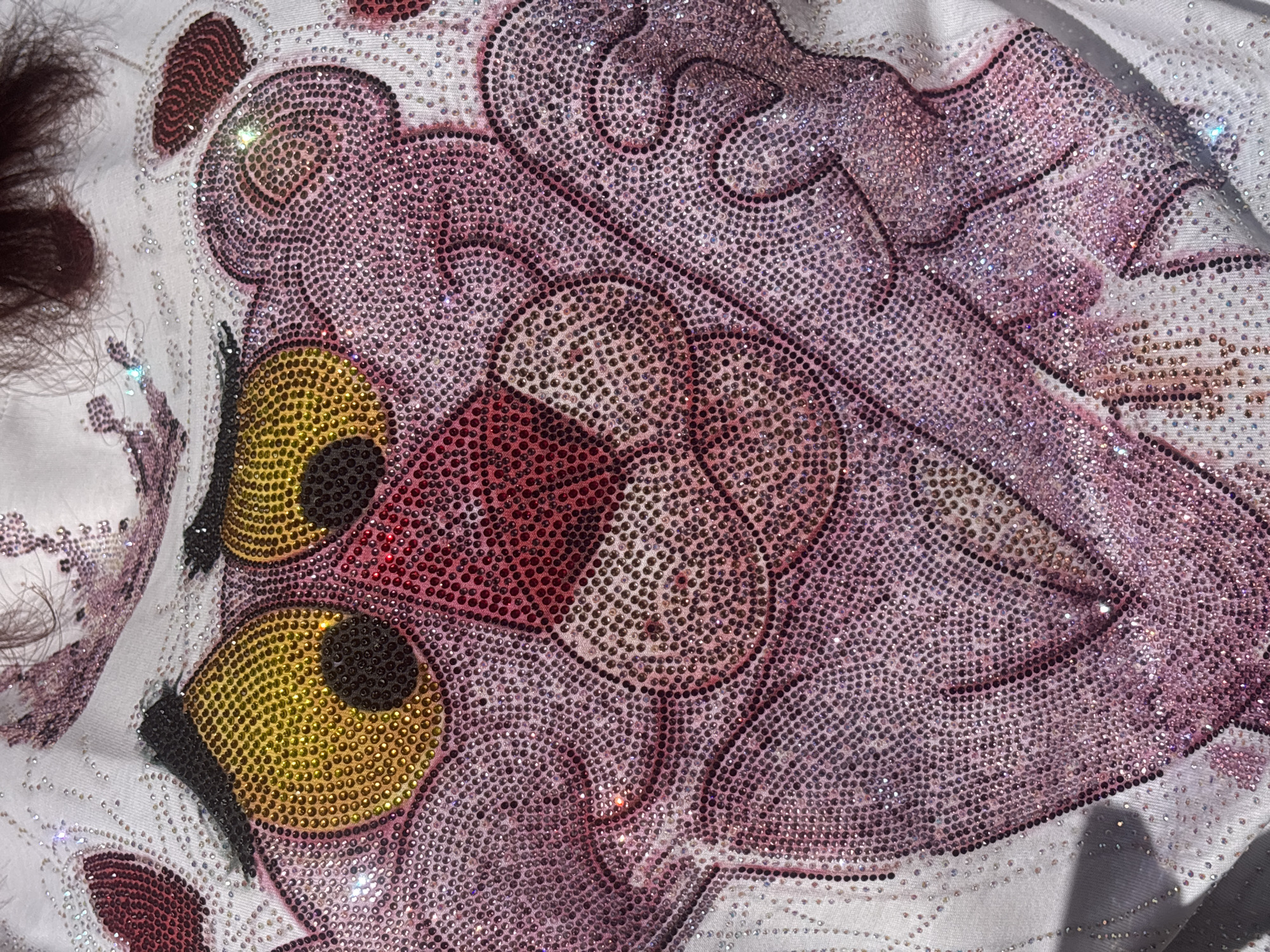 This close-up, high-angle shot depicts a textile item, likely clothing, intricately embellished with a rhinestone design. The central subject is a stylized cartoon character, strongly resembling the Pink Panther, rendered entirely in shimmering rhinestones. The character's body is composed of countless small rhinestones in various shades of pink and purple, catching the light and creating a sparkling effect. Its large, expressive eyes are a vibrant yellow, contrasted by solid black rhinestones forming the pupils and outlining certain features. A reddish-pink area marks the character's snout or nose. The rhinestones are precisely arranged to form distinct lines and filled shapes, contributing to the detailed texture of the design. The background is a soft, light-colored fabric, possibly white, against which the colorful embellishment stands out. On the far left side of the image, some dark, wispy fibers, resembling hair or fur, are visible. The bright lighting accentuates the individual facets of the rhinestones, creating numerous glints and highlights. No specific activity or event is taking place, as the image focuses on the static details of the embellished fabric. There is no visible text. The context for this image is Sofia, Bulgaria.