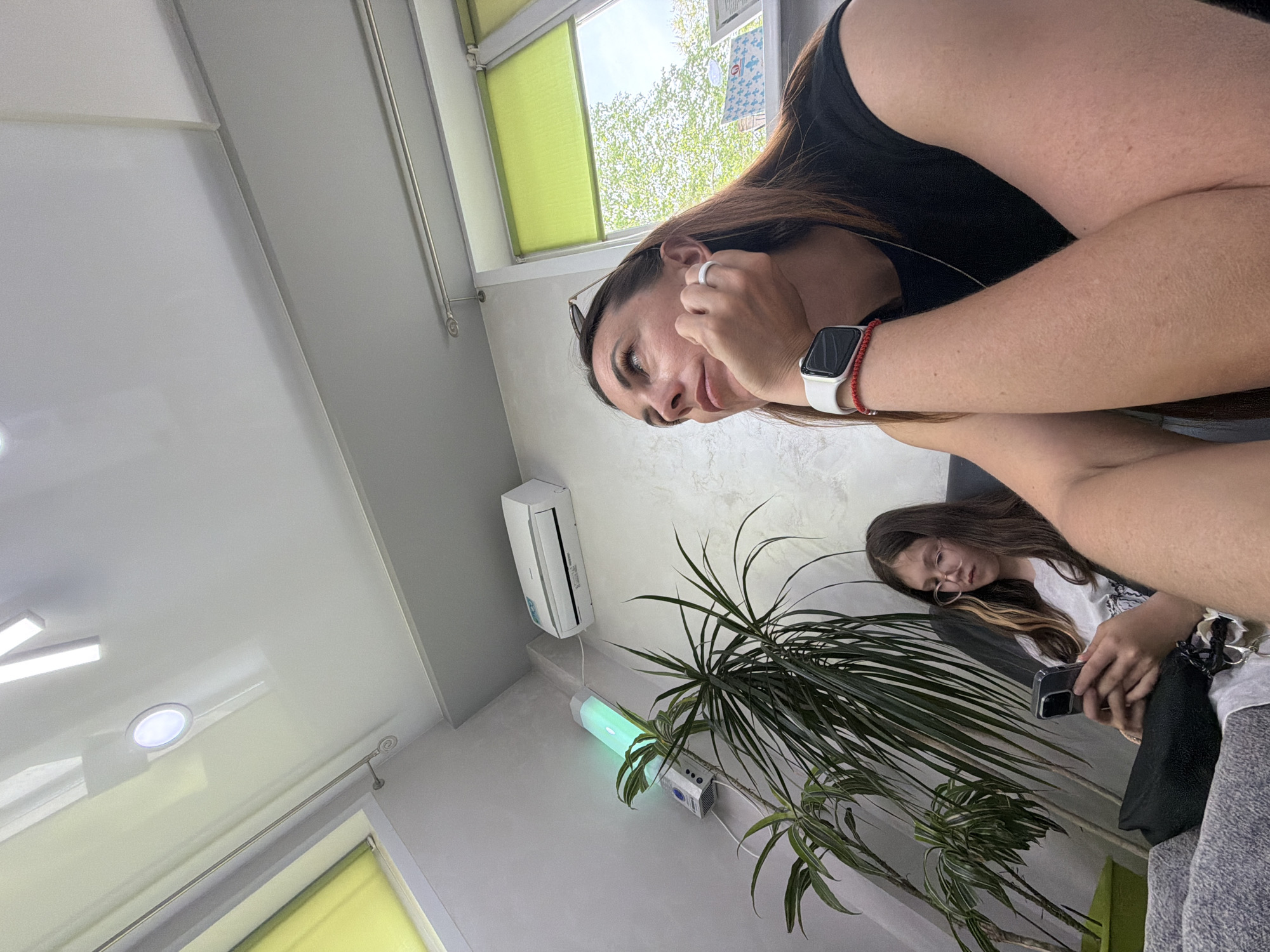 The image presents an indoor scene with two females and various objects in view. One female is in the foreground, partially obscuring the scene with her right arm raised towards her face. She has dark hair and is wearing a black tank top and an Apple Watch. The second female is in the background, focused on a smartphone, wearing glasses and a white t-shirt.
Environmentally, the location contains green window shades, an air conditioner unit mounted on the wall, a potted plant, a light fixture, and a white ceiling. The image appears to be taken indoors during daylight hours. No information indicates that the location is in the city of Birobidzhan, Russia.