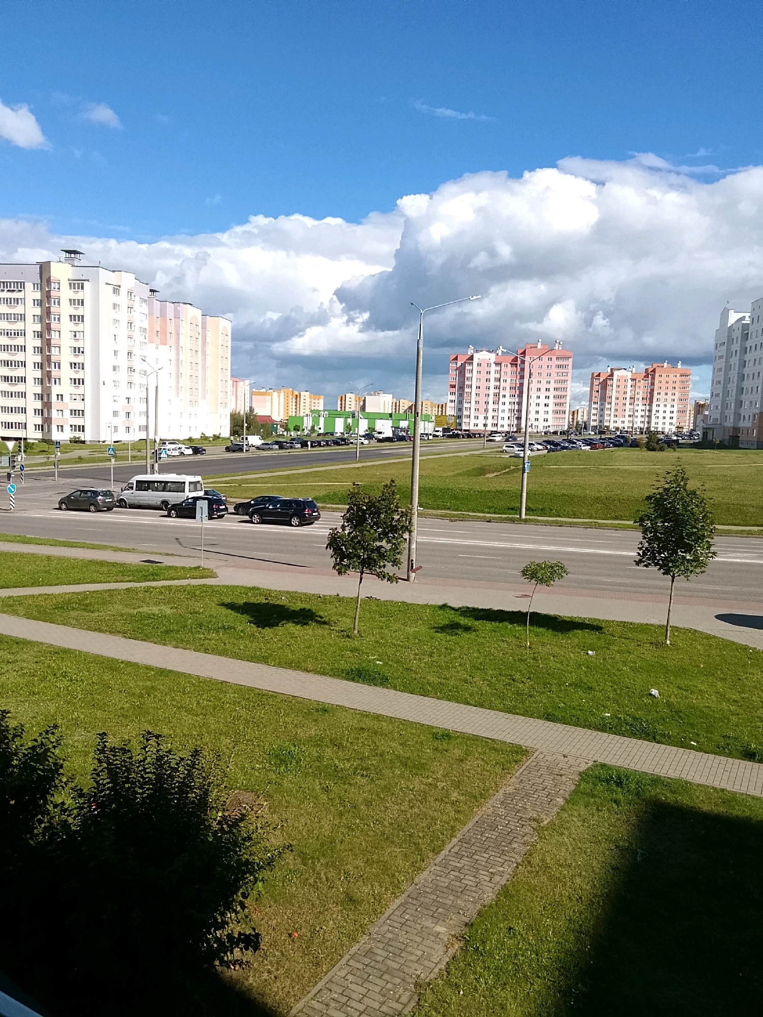 The image captures a contemporary urban landscape in Hrodna, Belarus, under a partly cloudy sky. In the background, several multi-story apartment buildings dominate the skyline. On the left, a pale yellow and white building features numerous windows and balconies. To its right, a series of buildings display facades in various pastel colors, including pink, peach, and beige, also with many windows. Further to the right, a white building stands out, and in the mid-ground, more apartment blocks in orange hues are visible.
A wide, asphalt road runs horizontally across the middle ground of the image, featuring multiple lanes and traffic markings. Several vehicles are present on the road, including a white van, a dark SUV, and other cars. A pedestrian crossing with traffic signals is visible on the left side of the road.
The foreground and areas adjacent to the road are covered in lush green grass. Paved walkways, constructed with interlocking bricks, crisscross the grassy areas. Several small trees are sparsely planted on the grass, with one larger tree casting a shadow in the lower right quadrant of the image. The overall scene suggests a well-maintained, residential area with modern infrastructure.