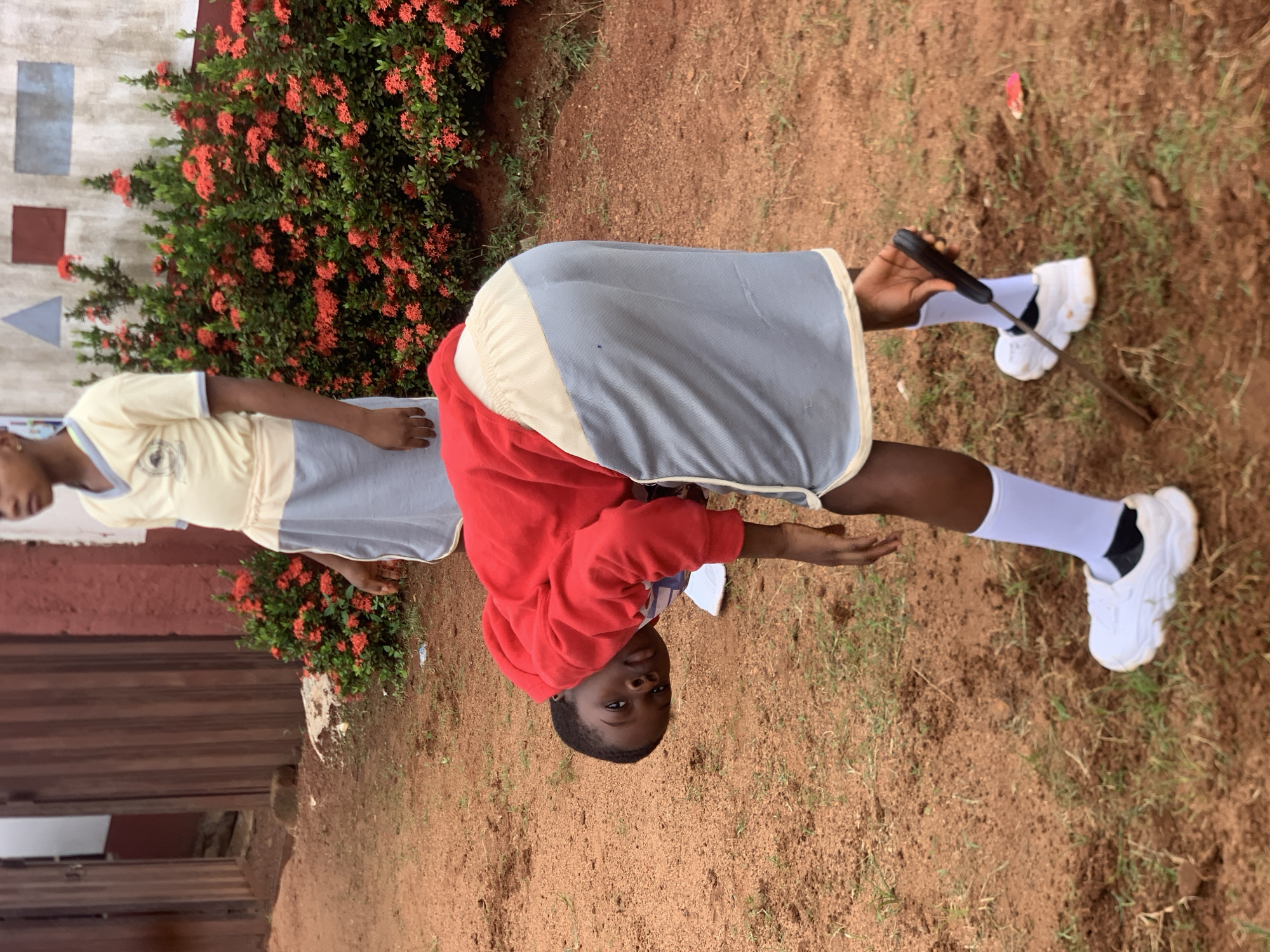The image depicts a low-angle outdoor scene in Ukwu-Abwa, Nigeria. Two children are visible, both wearing school uniforms consisting of a yellow shirt and a light grey skirt. One child, in the foreground, is bent over and appears to be digging or playing with the soil, wearing a red hoodie over their uniform and white socks with white sneakers. The other child, in the background, stands upright and observes. The environment features red earth, sparse green grass, and a dense bush with small red flowers. A wooden fence and a building with a reddish-brown wall are also present in the background. The overall lighting suggests an overcast or shaded day.