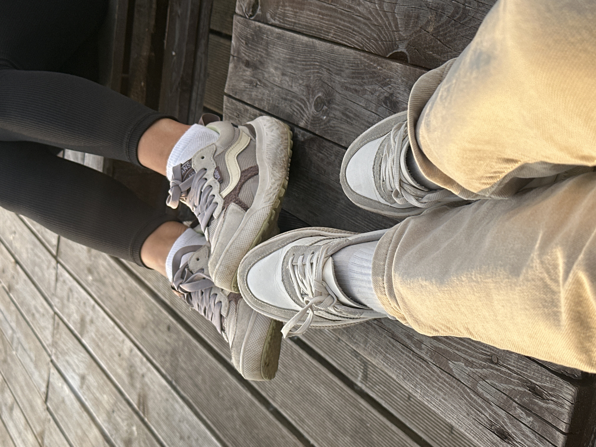 The image displays a low-angle perspective of two individuals' lower legs and feet, seated on a weathered wooden surface, purportedly in the city of Chaikovsky, Russia. In the immediate foreground, the first person wears light beige, possibly corduroy, pants with cuffed hems, exposing white ribbed ankle socks and light-colored (white and light grey) laced sneakers.
Further back in the mid-ground, the second person is depicted wearing dark grey or black ribbed leggings, paired with white ankle socks and beige and grey Vans brand sneakers. The Vans logo, including the sidestripe and a small tag, is visible on the side of the shoes, which feature a chunky, textured, and slightly soiled sole.
Both individuals' feet are positioned on a greyish-brown wooden platform or decking, composed of planks with visible grain patterns, knots, and gaps. Some planks show signs of wear and dirt. A slightly raised wooden edge or step is discernible beneath the mid-ground person's legs. The scene is illuminated by warm, directional light, suggesting late afternoon or early morning conditions, which highlights the textures of the clothing, footwear, and wood.