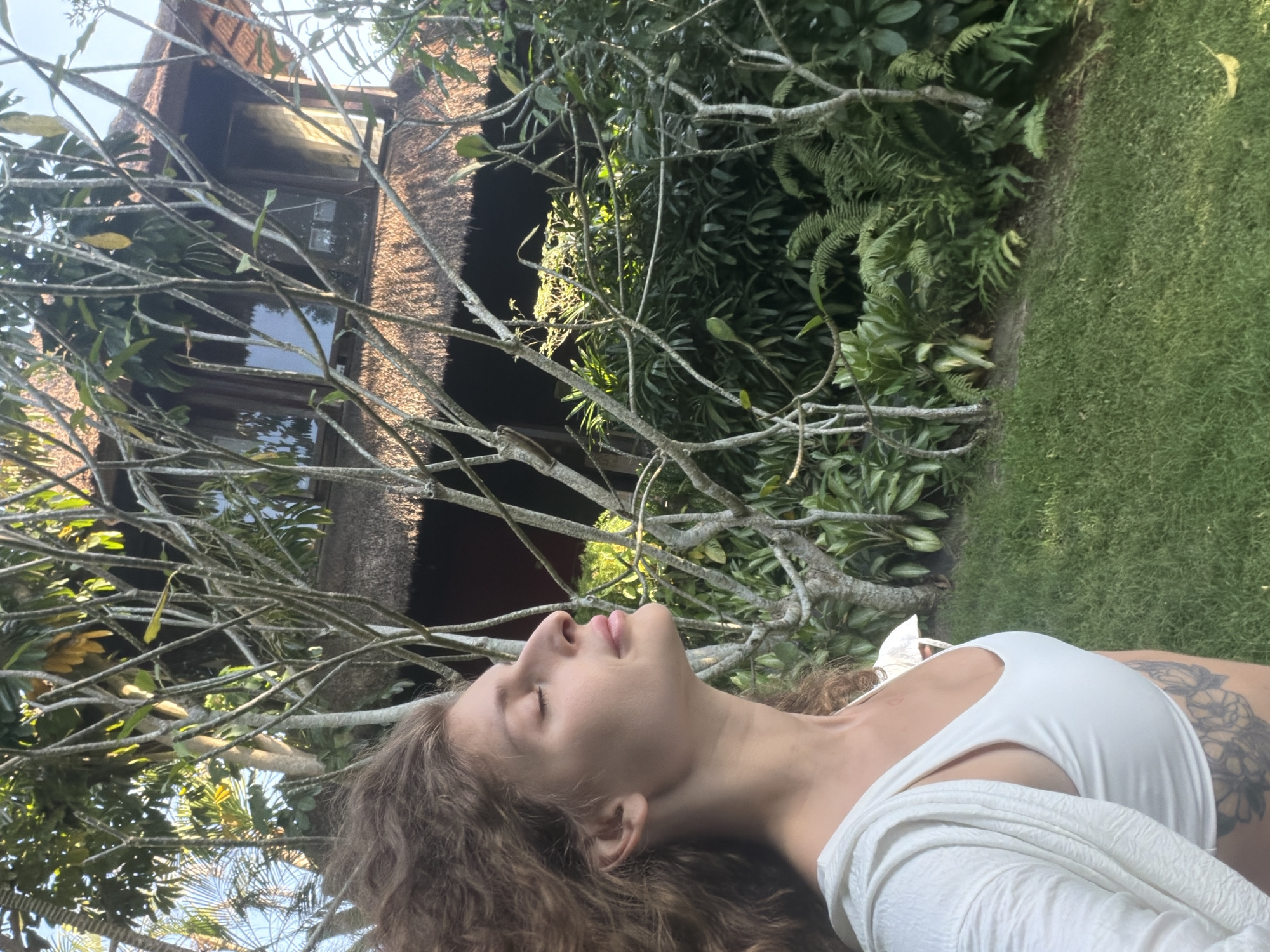 The image captures a woman positioned on the left side of the frame, facing slightly to her right with her eyes closed and head tilted upwards, appearing to experience her surroundings. Her mid-length, curly brown hair is visibly wind-blown, sweeping backwards to her left. She wears a white sports bra or crop top beneath an open, textured white long-sleeved cover-up. A floral tattoo is visible on her lower right torso, specifically on her rib cage area.
The background features a lush tropical environment, consistent with the specified location of Mengwi, Indonesia. A traditional structure, likely a house or villa, is partially visible in the upper right. It features a distinct dark brown thatched roof and wooden elements, including two visible windows on an upper story that reflect the bright sky. Dense, predominantly green foliage fills the midground and background, including large-leafed plants, ferns, and numerous thinner, light-barked trees with sparse leaves that stand between the viewer and the background structure. A well-maintained lawn with short green grass occupies the lower portion of the image, sloping gently upwards towards the vegetation. One yellow leaf rests on the grass in the lower right. Bright, natural sunlight illuminates the scene from the right, casting soft shadows and highlighting the woman's profile and the vibrant greenery. The sky, a clear light blue, is visible in the upper left corner through gaps in the foliage.