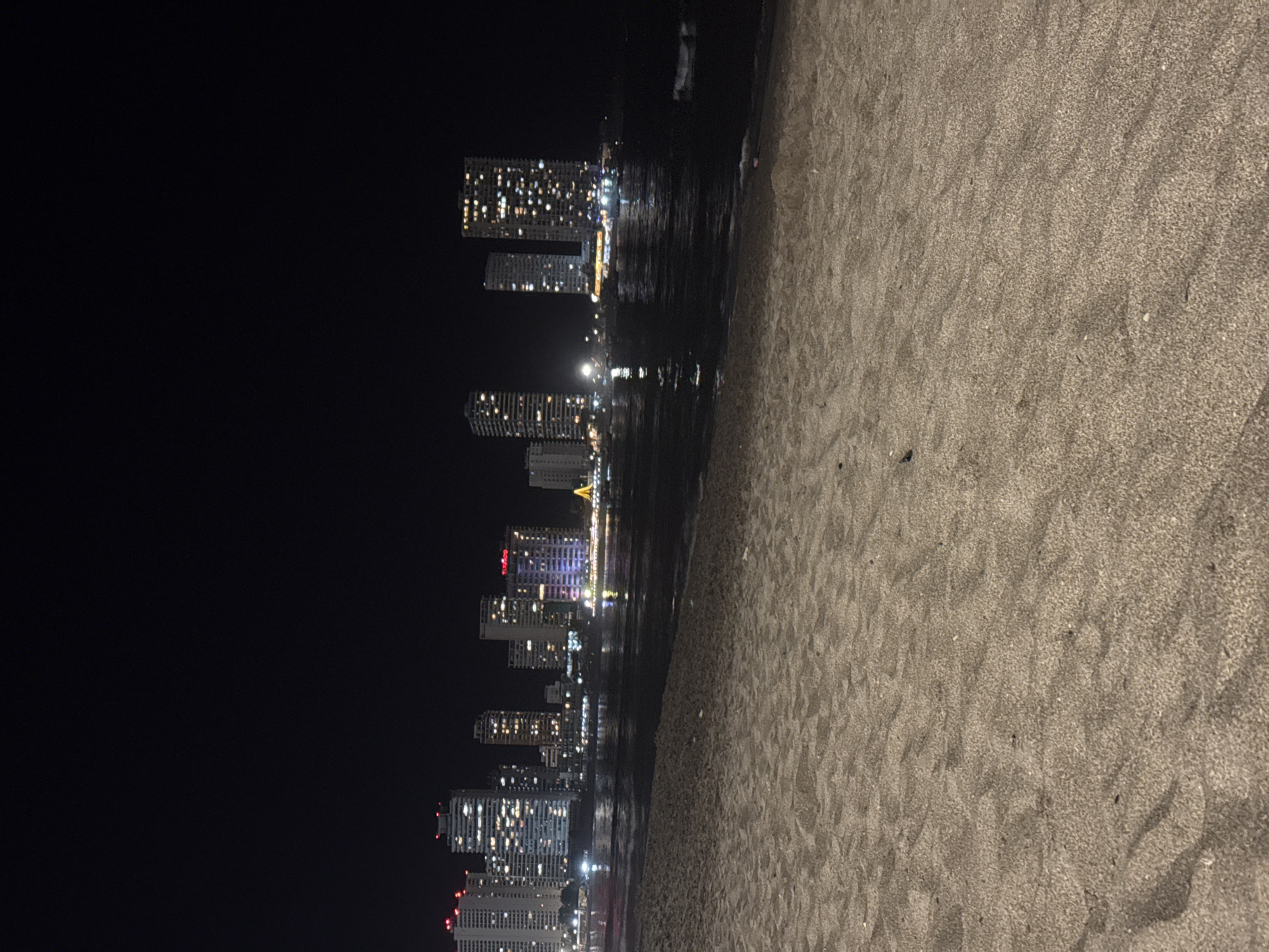 At night, a coastal scene depicts a beach with a dark sea and city skyline. The beach is composed of gray sand. The city skyline includes several tall buildings with illuminated windows. The sea appears dark, reflecting the city lights. The scene is illuminated by city lights.