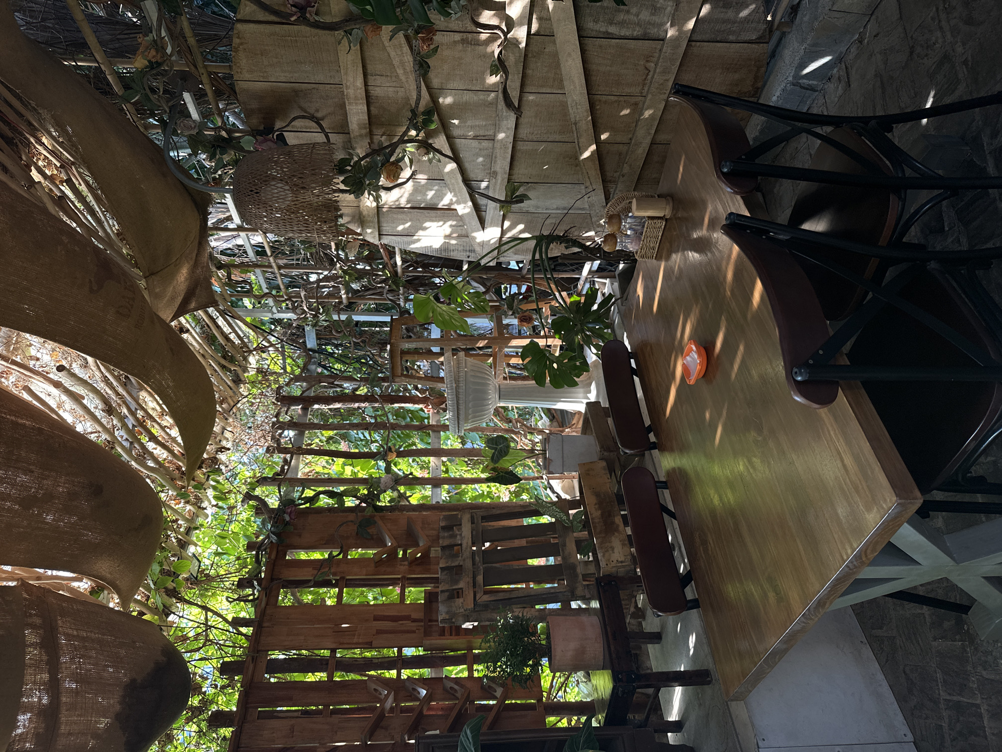 The image shows an outdoor seating area with a wooden table and chairs. The area is covered by a rustic canopy of burlap and branches, with vines and plants growing around it. A wooden fence is visible behind the table, and a large wooden barrel-like structure is to the right. The table is set with salt and pepper shakers and a small orange dish. The lighting suggests it is daytime, with sunlight filtering through the canopy and casting shadows. The scene appears to be in a restaurant or cafe with a natural, bohemian aesthetic. While the specific location in Nha Trang Ward, Vietnam is not visually evident from the image alone, the description implies a natural, possibly shaded outdoor space in a restaurant setting.