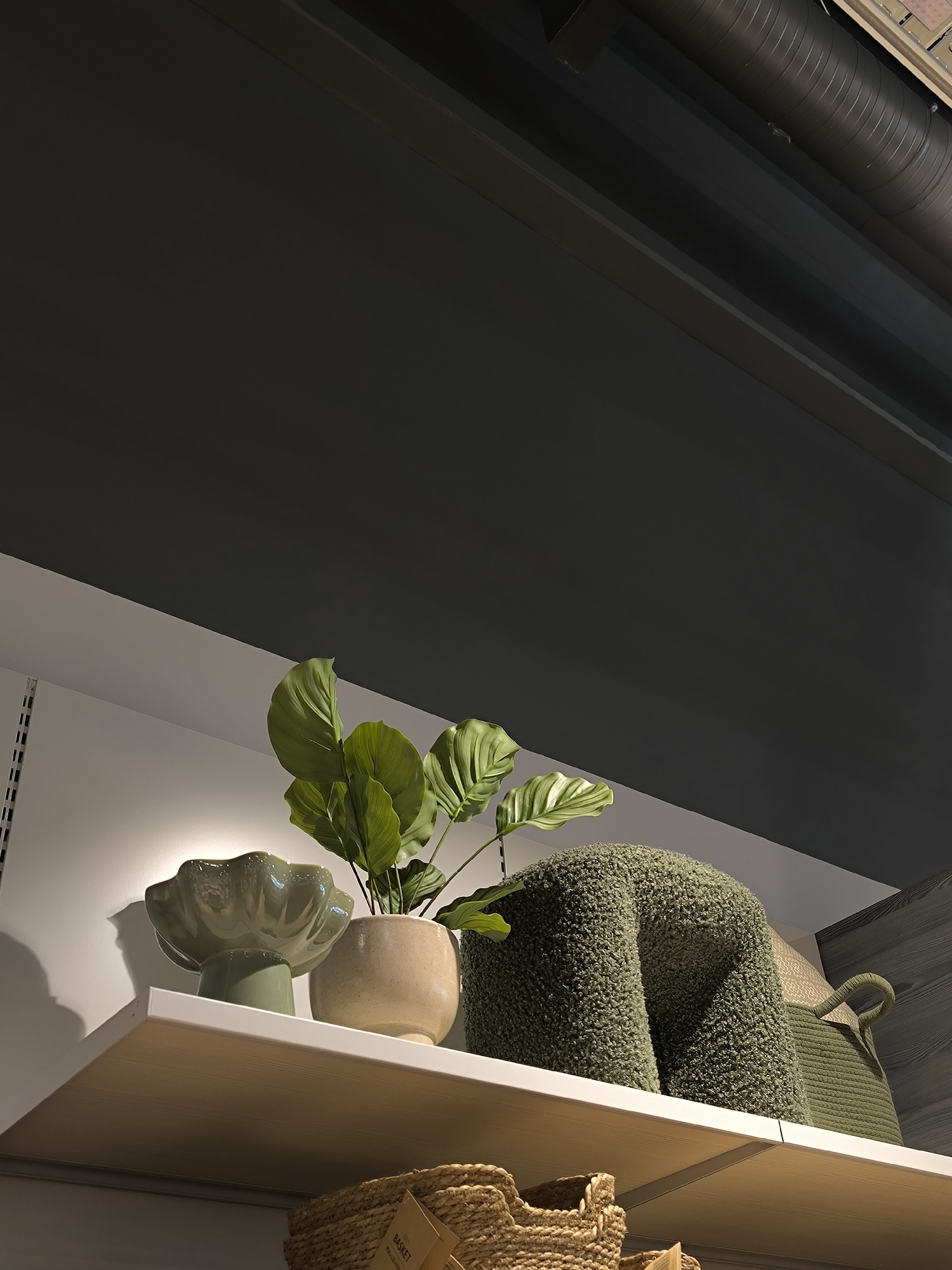The image captures a low-angle view of a white shelf adorned with various decorative items, set against a two-toned wall. The upper portion of the wall, along with the ceiling, is a dark charcoal grey, while a narrow white strip separates it from a lighter, off-white wall behind the shelf. In the upper right, a dark, corrugated pipe or ventilation duct is visible against the dark ceiling.
On the left side of the shelf, a metal shelving bracket with vertical slots is partially visible. The shelf itself holds several objects: a green, shell-shaped ceramic bowl or stand with a fluted edge; a vibrant green plant with large leaves in a light beige pot; and a textured, olive green object, possibly a cushion or sculpture, that appears to be folded or shaped like an inverted "U". To the right of these items, a woven green basket with a rounded handle is partially visible. The shelf is illuminated by a light source from above, casting subtle shadows behind the items on the back wall.
Below the main shelf, another, partially visible wicker basket is present. A small, light-colored label attached to this basket clearly shows the word "BASKET" in black text, with smaller, less legible text underneath it. The underside of the white shelf is a warm, light brown, suggesting a wood finish or a contrasting material. The overall impression is a modern, styled display, possibly within a retail environment or a contemporary home interior.