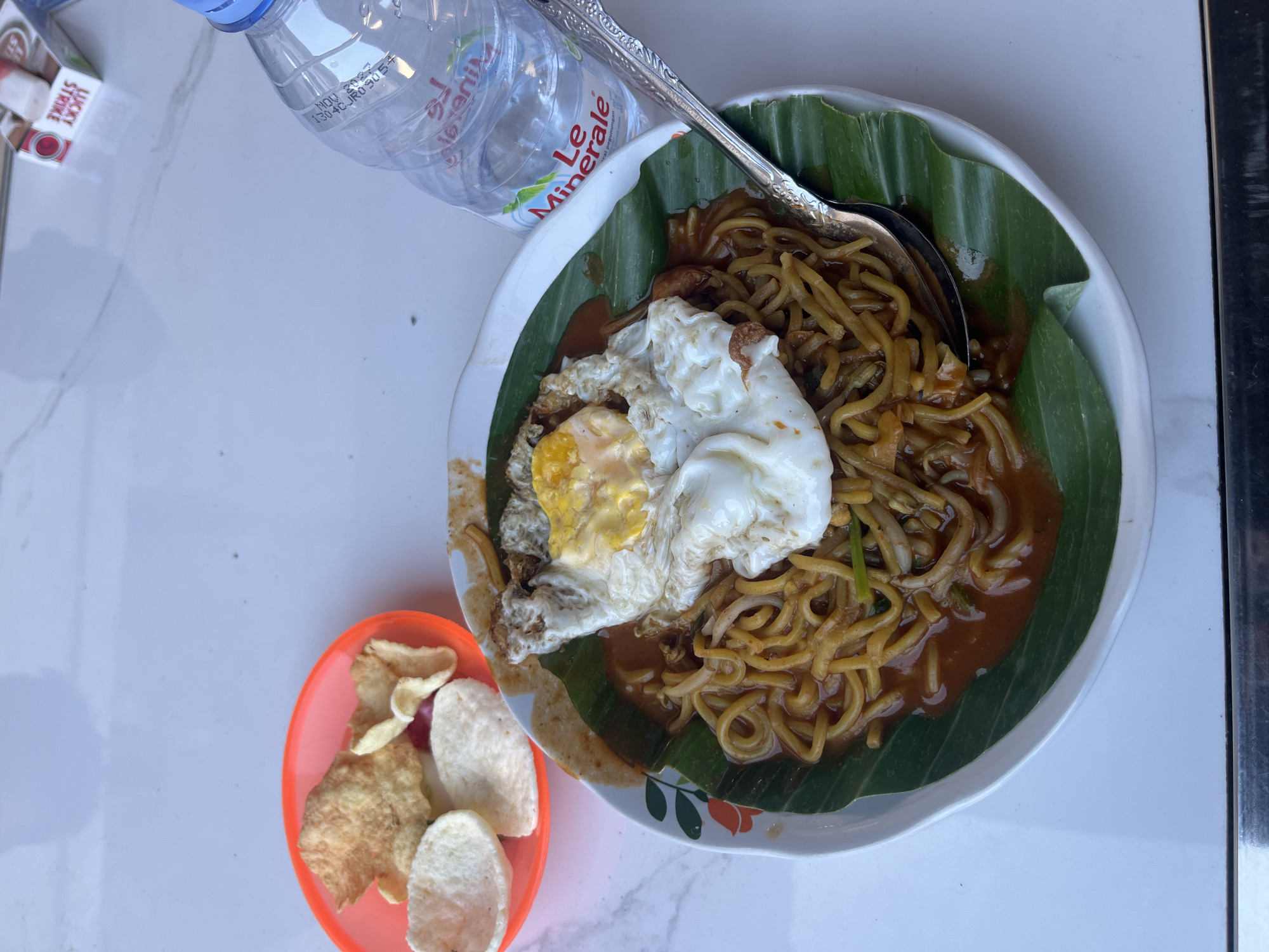 A plate of mie goreng, a savory Indonesian stir-fried noodle dish, is presented on a white plate lined with a banana leaf. The noodles are coated in a reddish-brown sauce and topped with a fried egg. To the left of the plate, a small orange dish contains a few pieces of crackers or krupuk. A bottle of "Le Minerale" drinking water is positioned to the right of the noodle plate. The scene appears to be set on a white, marbled surface, possibly a table. In the background, a partial view of a cigarette pack, identified as "Lucky Strike," is visible. The overall setting suggests an outdoor dining environment in Glameunasah Baro, Indonesia, where this meal is being served.