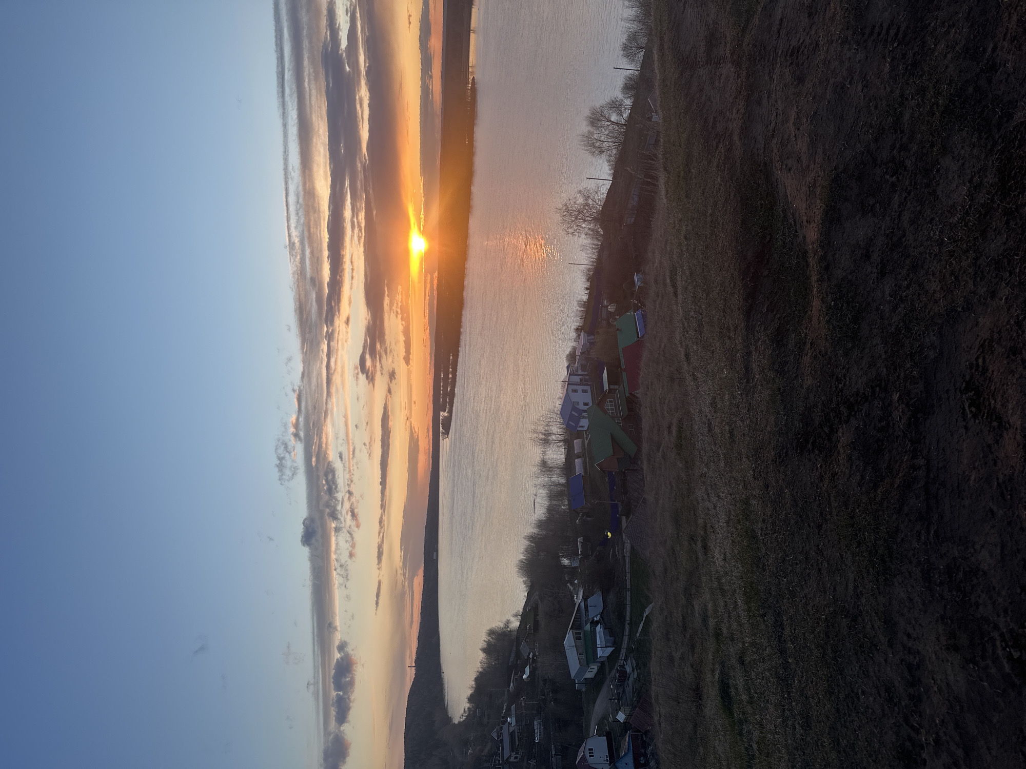 The image captures a sunset over a body of water, likely a river, in Ольховка, Russia. The sun is positioned on the horizon, reflecting light on the water's surface. The sky is filled with clouds, ranging from light to dark, with the sunset casting an orange and yellow glow. On the right side of the image is a grassy slope leading down to a settlement with small houses featuring roofs of various colors, including blue, green, and red. Some trees are visible along the riverbank, their branches bare, suggesting it may be early spring or late fall. The composition suggests a vantage point from an elevated position overlooking the scene.