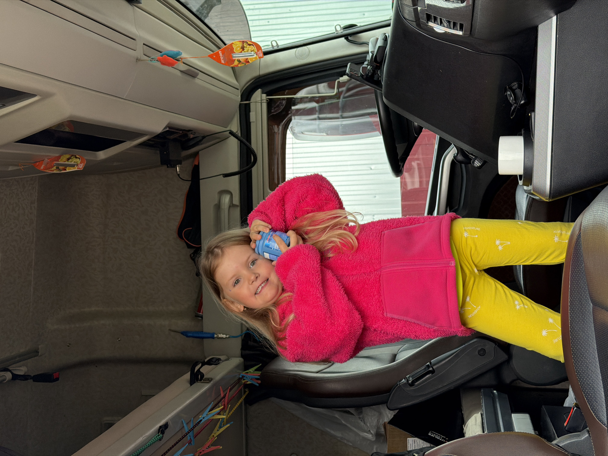 A young girl with blonde hair and blue eyes smiles brightly, looking directly at the camera. She is wearing a vibrant pink fuzzy jacket and yellow pants adorned with white palm tree motifs and the word "ADVENTURE." The girl is lying on her side on a grey and brown seat, holding a blue bottle that has "Macton" and "30" written on it.
The scene is set inside what appears to be the cabin of a large vehicle, possibly a truck or camper, evident from the interior layout, overhead compartments, and a partially visible steering wheel on the right. Daylight filters in through the windows, illuminating the interior. Various personal items and accessories are visible: several air fresheners, including one shaped like a leaf with the brand "AREON" and images of fruit, and two smaller, colorful, mouse-shaped ones. Cables and wires are strung across different surfaces, and a line with colorful plastic clothes pegs can be seen on the left, suggesting a lived-in space. The girl's joyful expression suggests a relaxed and happy atmosphere. The location is Kamyennoye, Russia.