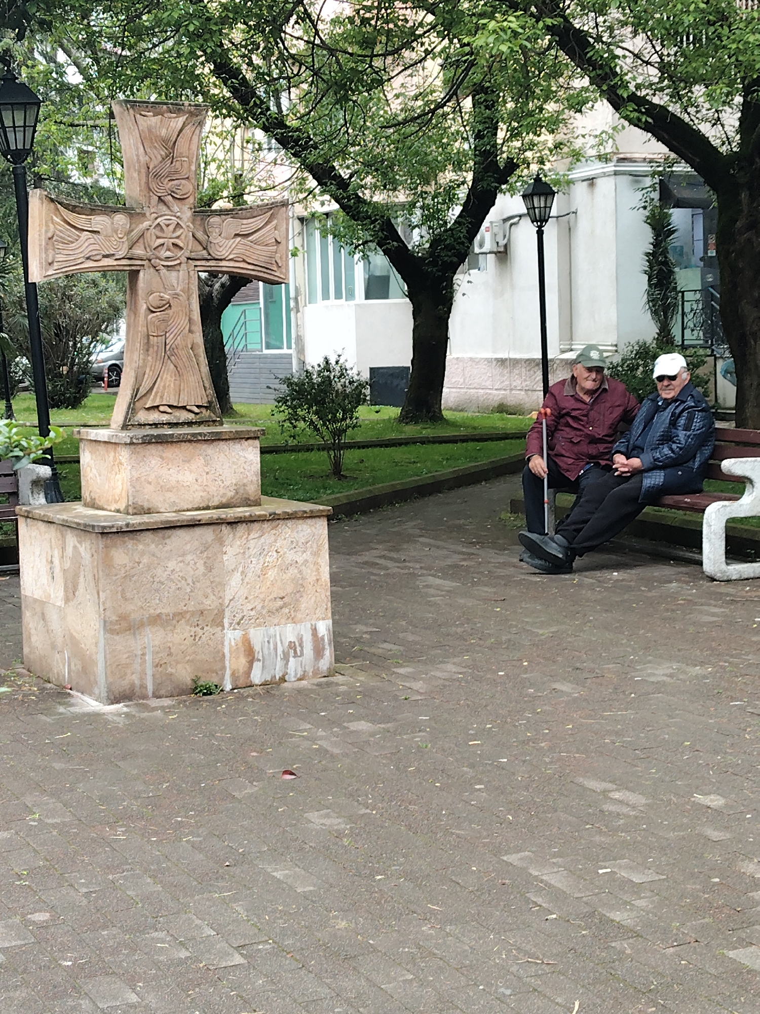 In a city park in Batumi, Georgia, a large stone cross stands on a square base on the left, while two elderly men are seated on a bench on the right. The cross is intricately carved with religious symbols. The men are dressed in casual attire, with one holding a cane. Trees and a building are visible in the background. The ground is paved with interlocking stones.